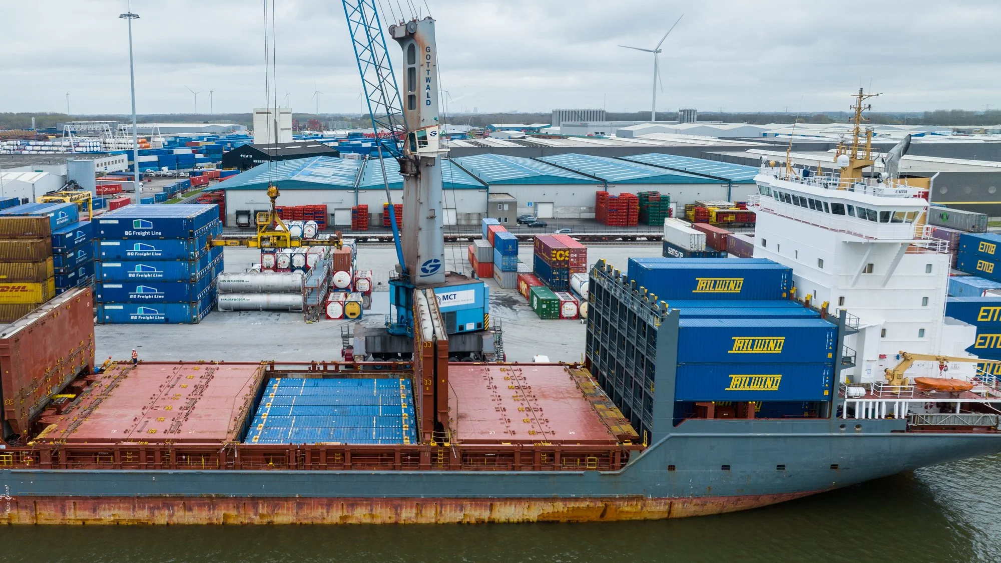 Cargo ship docked at a port with containers, a crane, and a warehouse in the background, under an overcast sky.