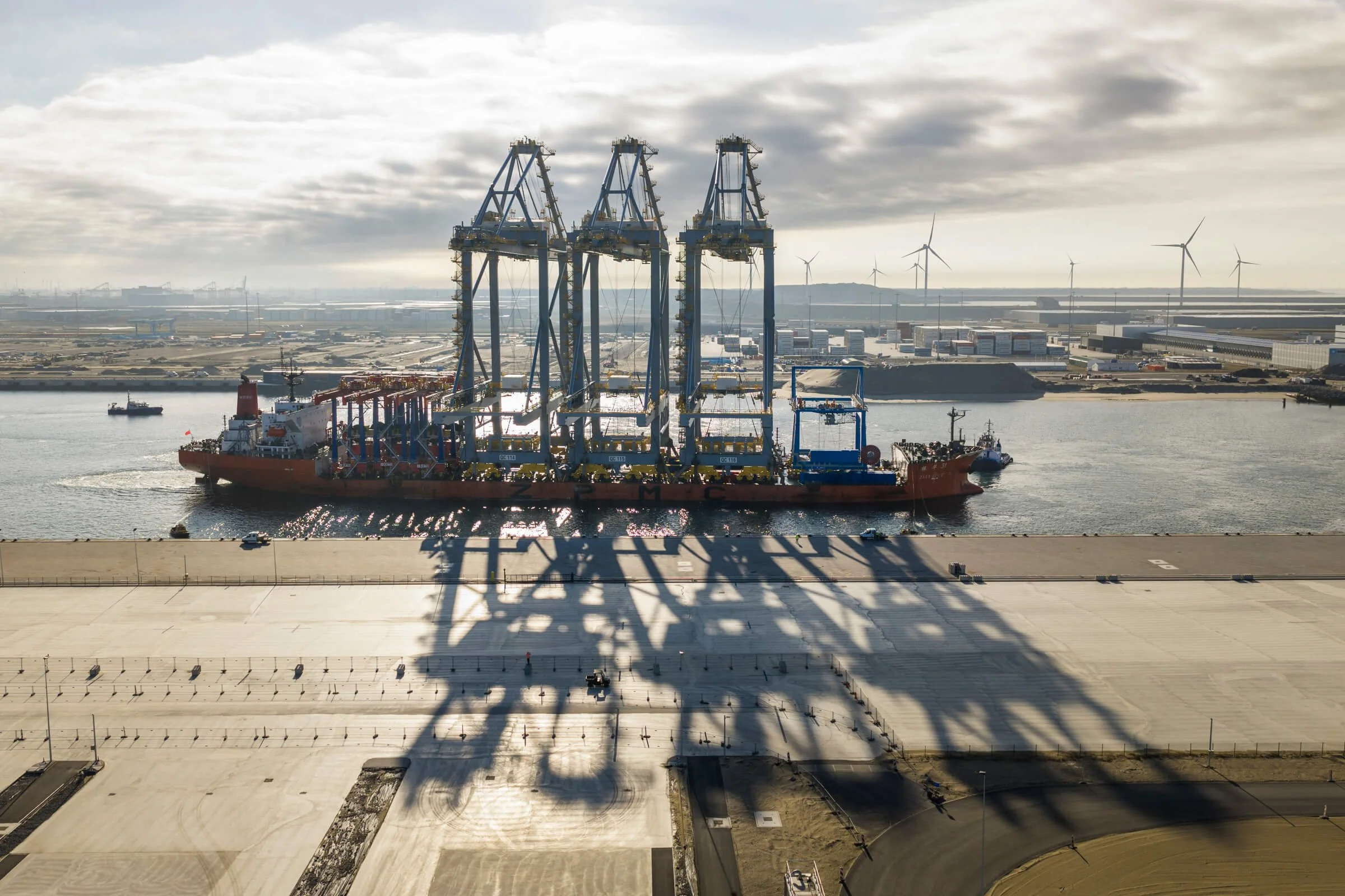 Large cargo ship with blue cranes in a harbor, shadowed on the concrete ground, with wind turbines in the background and cloudy sky.