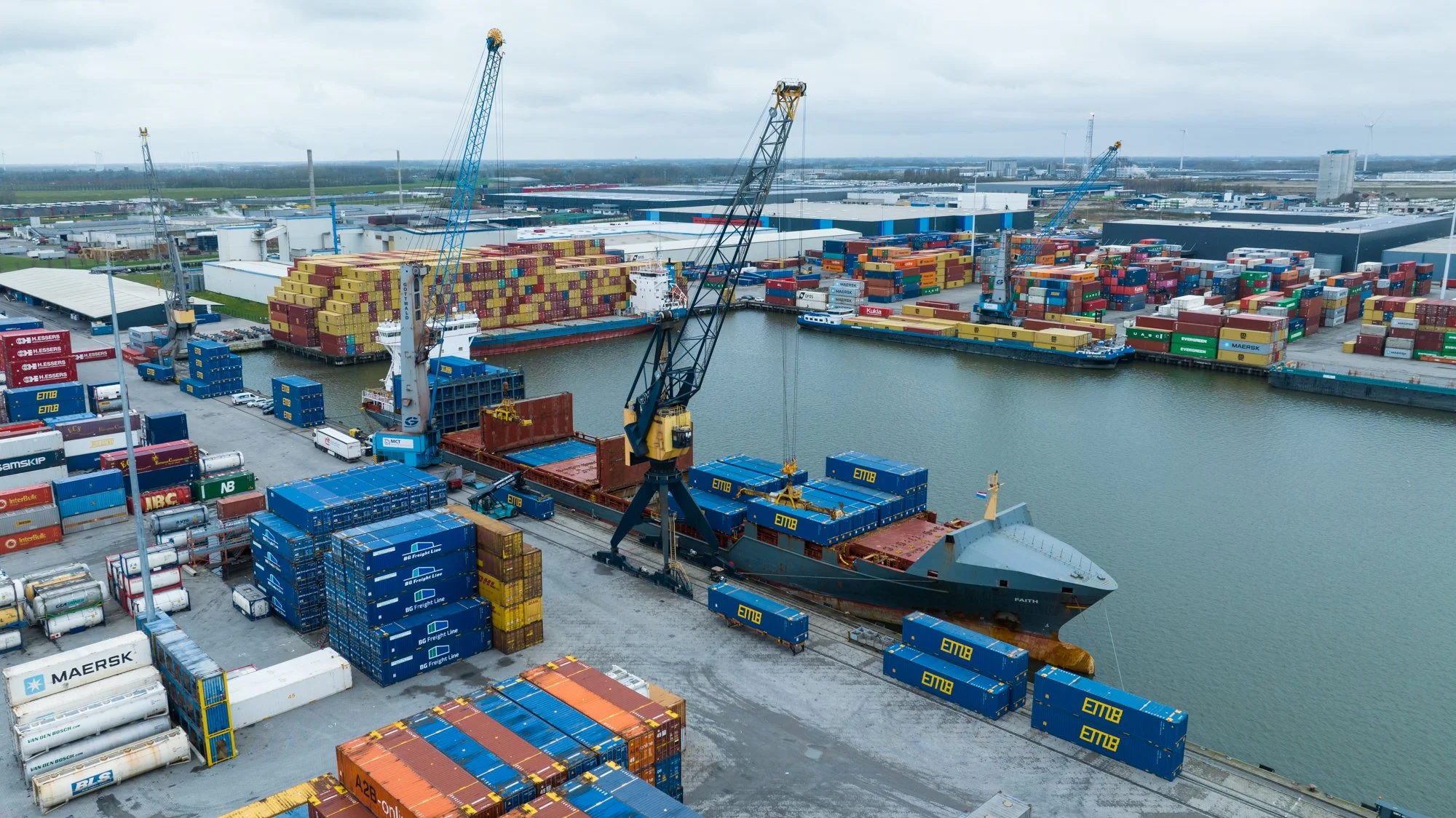 A busy shipping port with large cranes unloading cargo containers from ships onto the dock, with numerous colorful containers stacked throughout the area, and water surrounding the port.