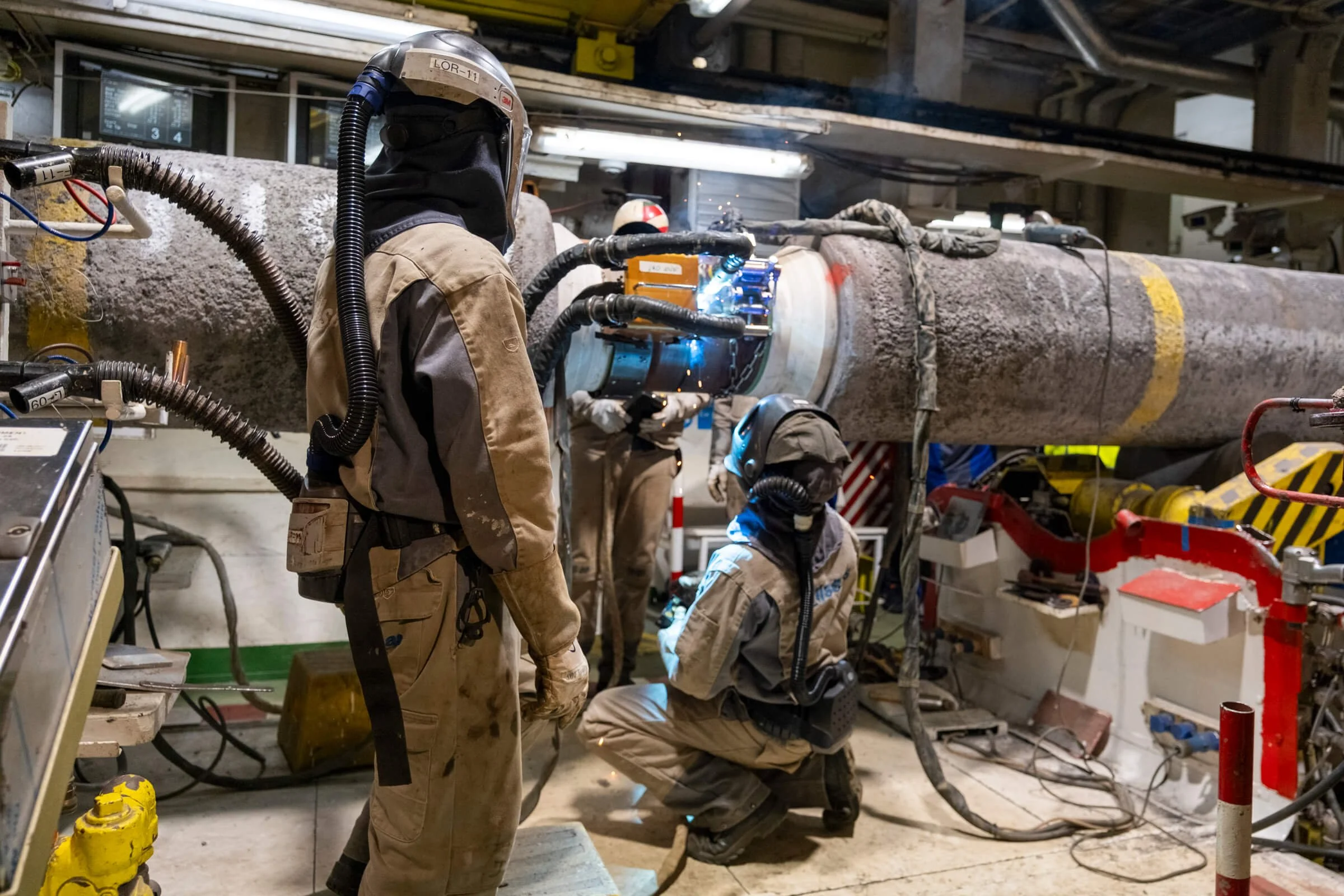 Three workers in protective gear welding a large pipeline in an industrial setting.