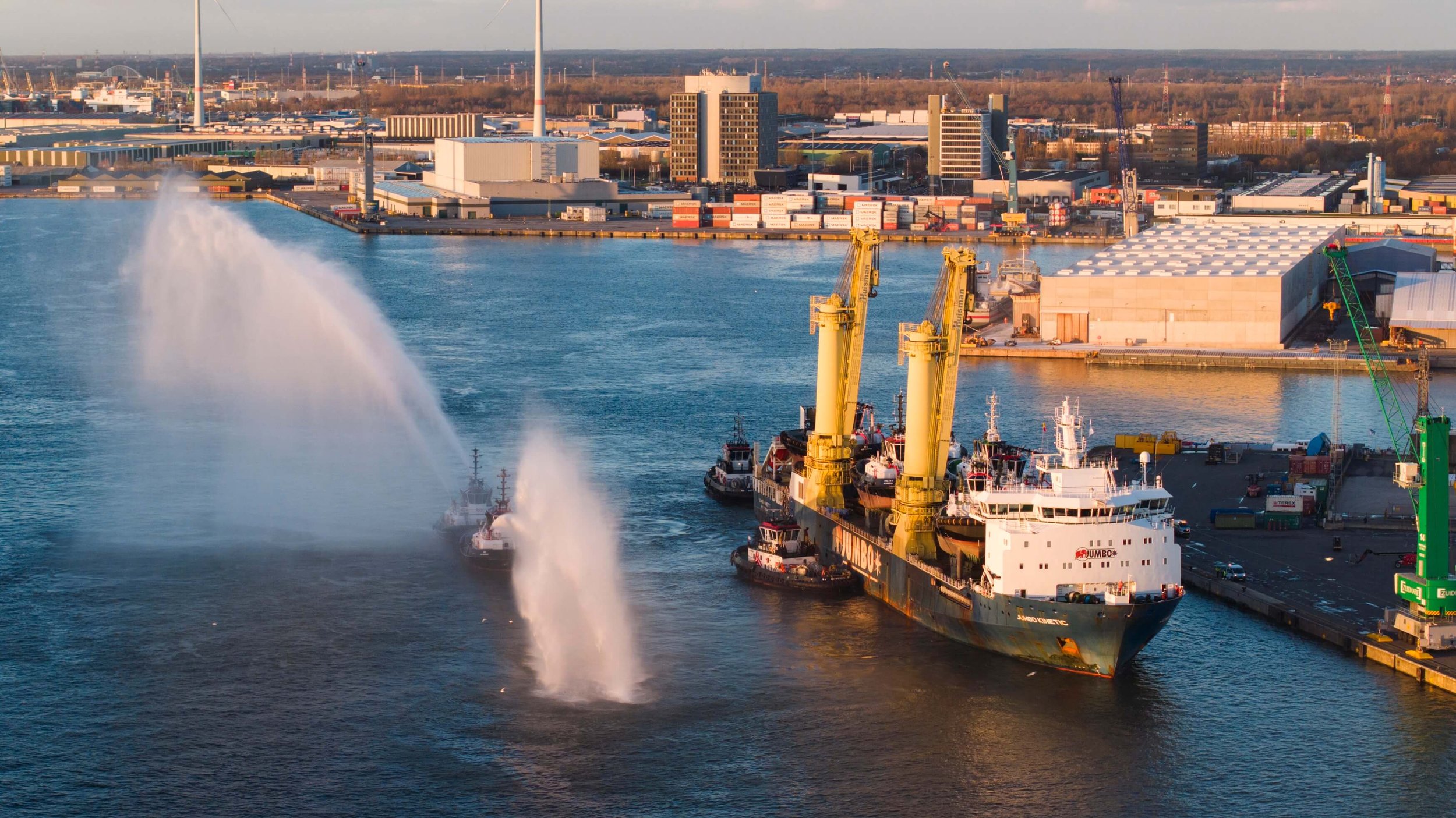 A harbor with a large ship docked, three tugboats spraying water, industrial warehouses, cranes, and city buildings in the background.