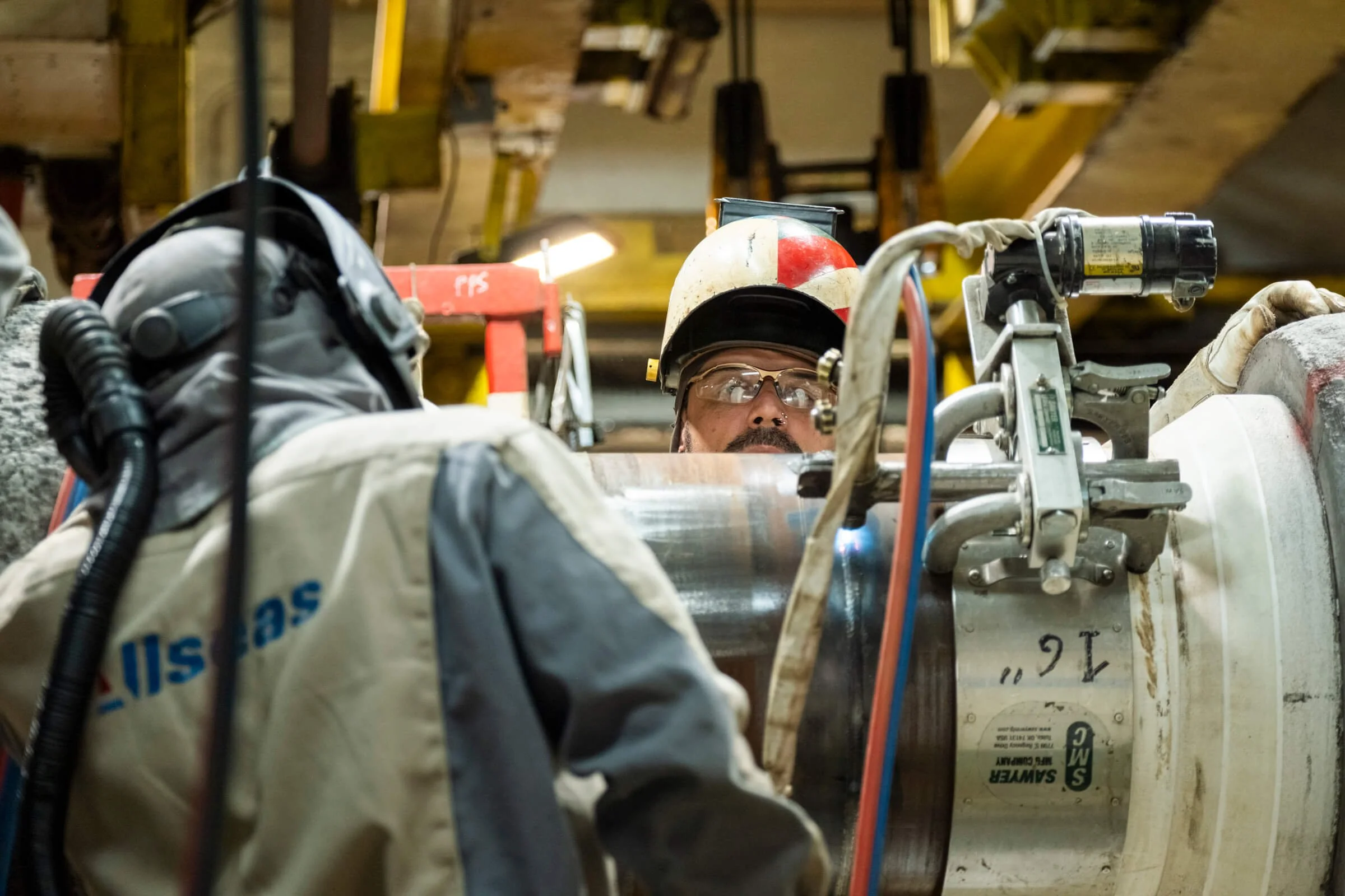 A person in a helmet and protective glasses working on industrial machinery in a workshop, with wooden beams and tools visible in the background.