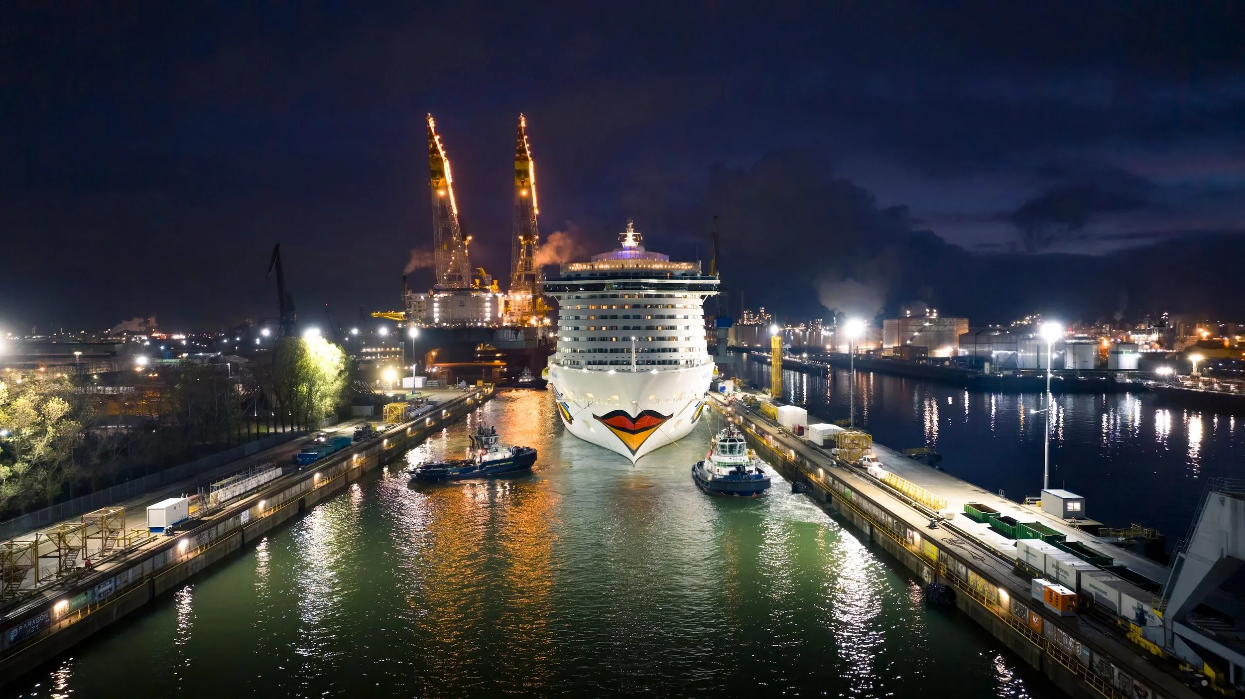 A cruise ship with a lipstick kiss symbol on the bow is docked in a harbor at night, lit by numerous artificial lights with industrial structures and cranes in the background.