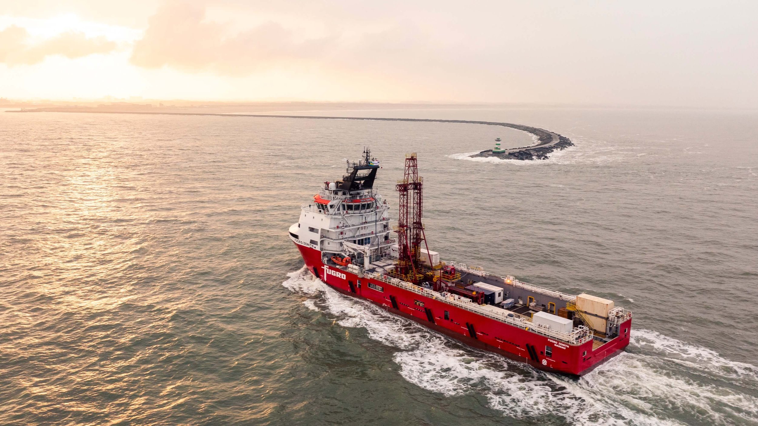A large red and white vessel sailing in the ocean, with a cityscape visible in the distance and a curved breakwater extending into the water.
