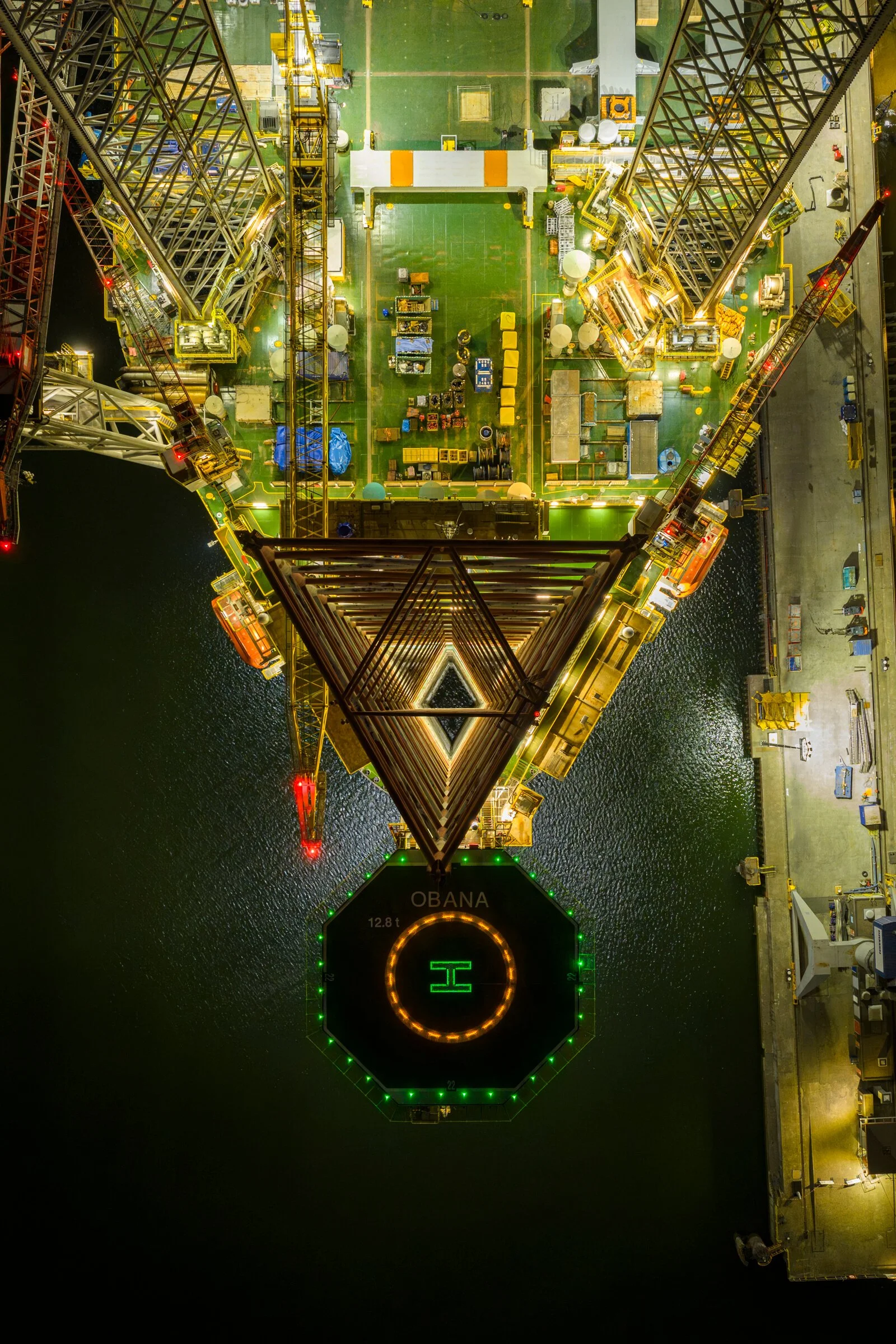Nighttime aerial view of a tall oil platform in the ocean, illuminated with yellow, green, and orange lights, with water visible around it.