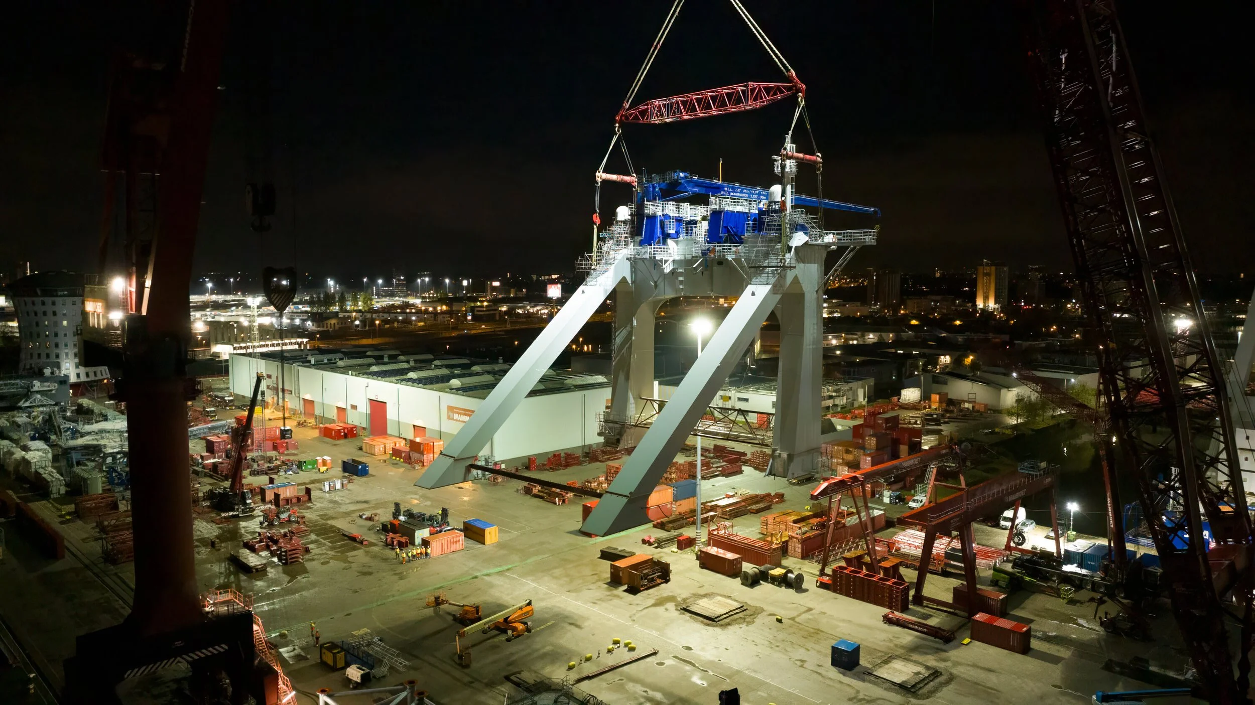 Nighttime scene of a large construction site with a massive ship being built or assembled, surrounded by equipment, cranes, and containers, with city lights visible in the background.