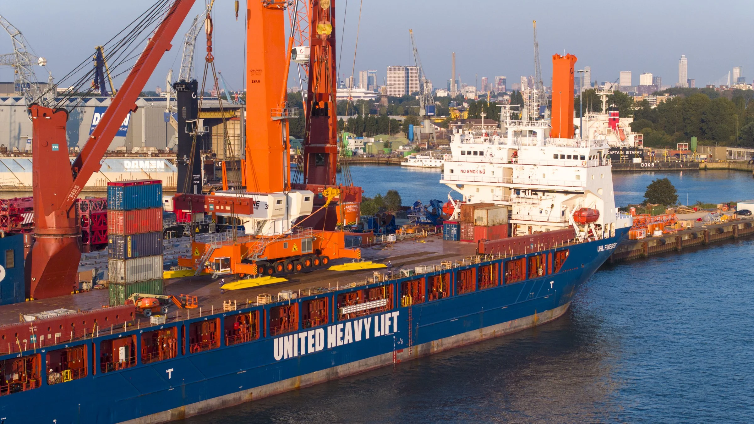A large cargo ship named 'United Heavy Lift' docked at a busy port with cranes, containers, and a city skyline in the background.