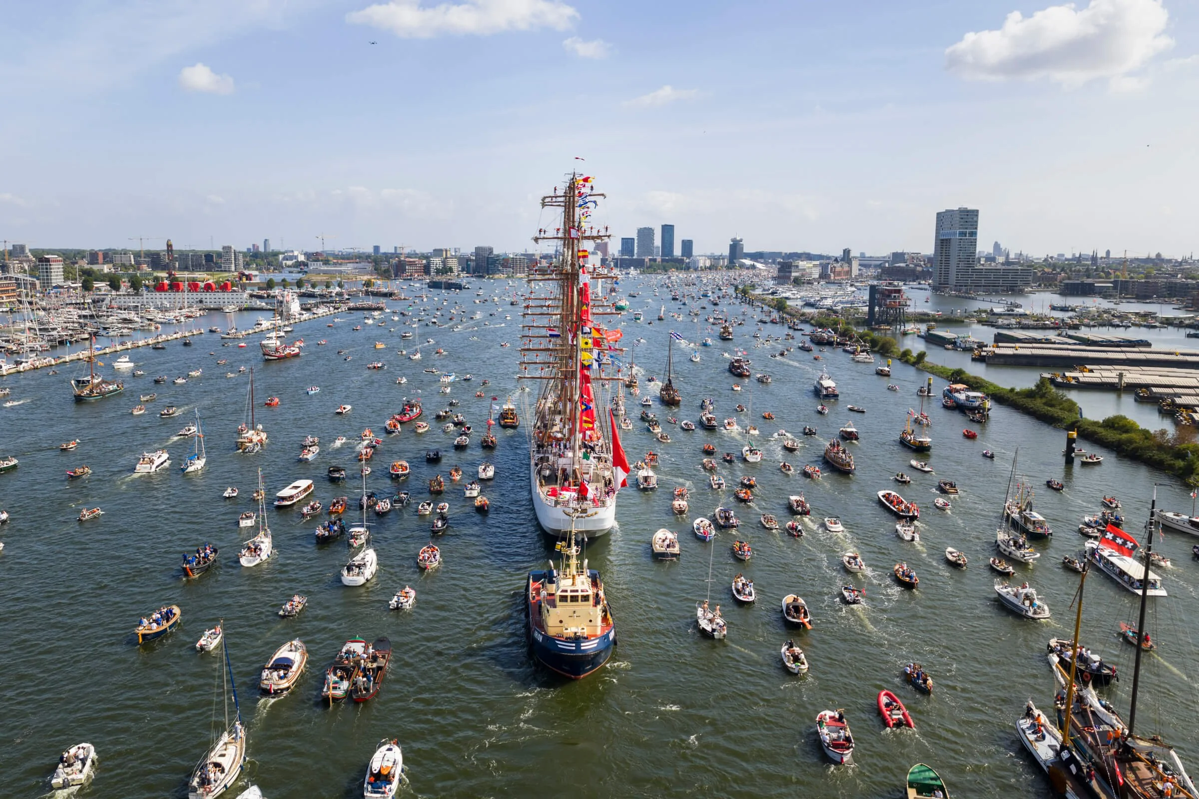Large white sailing ship with colorful flags, surrounded by many smaller boats in a city harbor with modern buildings and a cloudy sky.