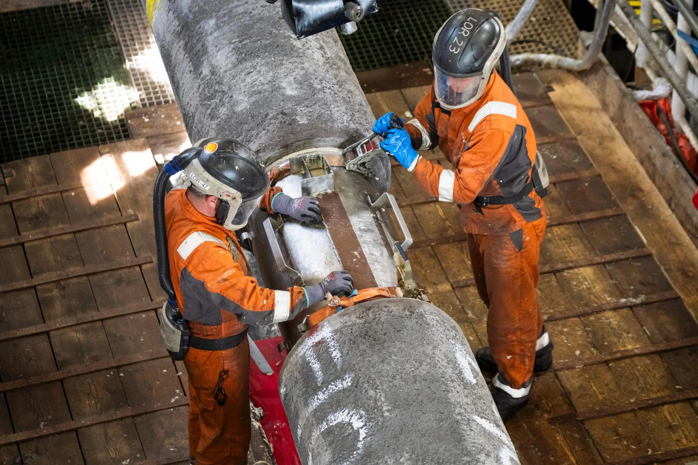 Two workers in orange safety suits, helmets, and gloves are inspecting and working on a large pipeline gauge. They are surrounded by wooden flooring and industrial equipment.