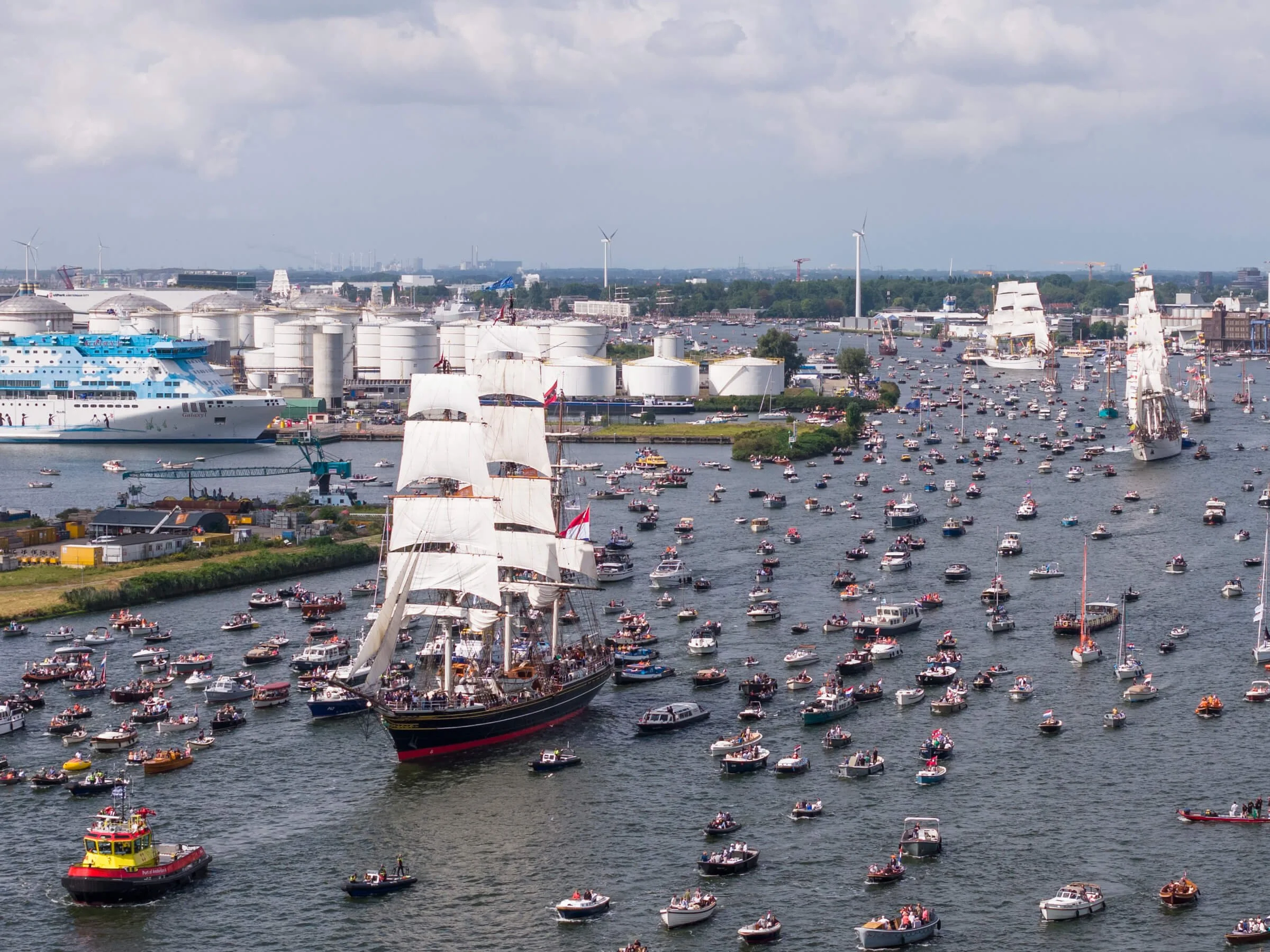 A harbor scene with numerous boats and ships, including a large sailing ship with white sails and a colorful passenger boat, surrounded by industrial buildings and wind turbines in the background.