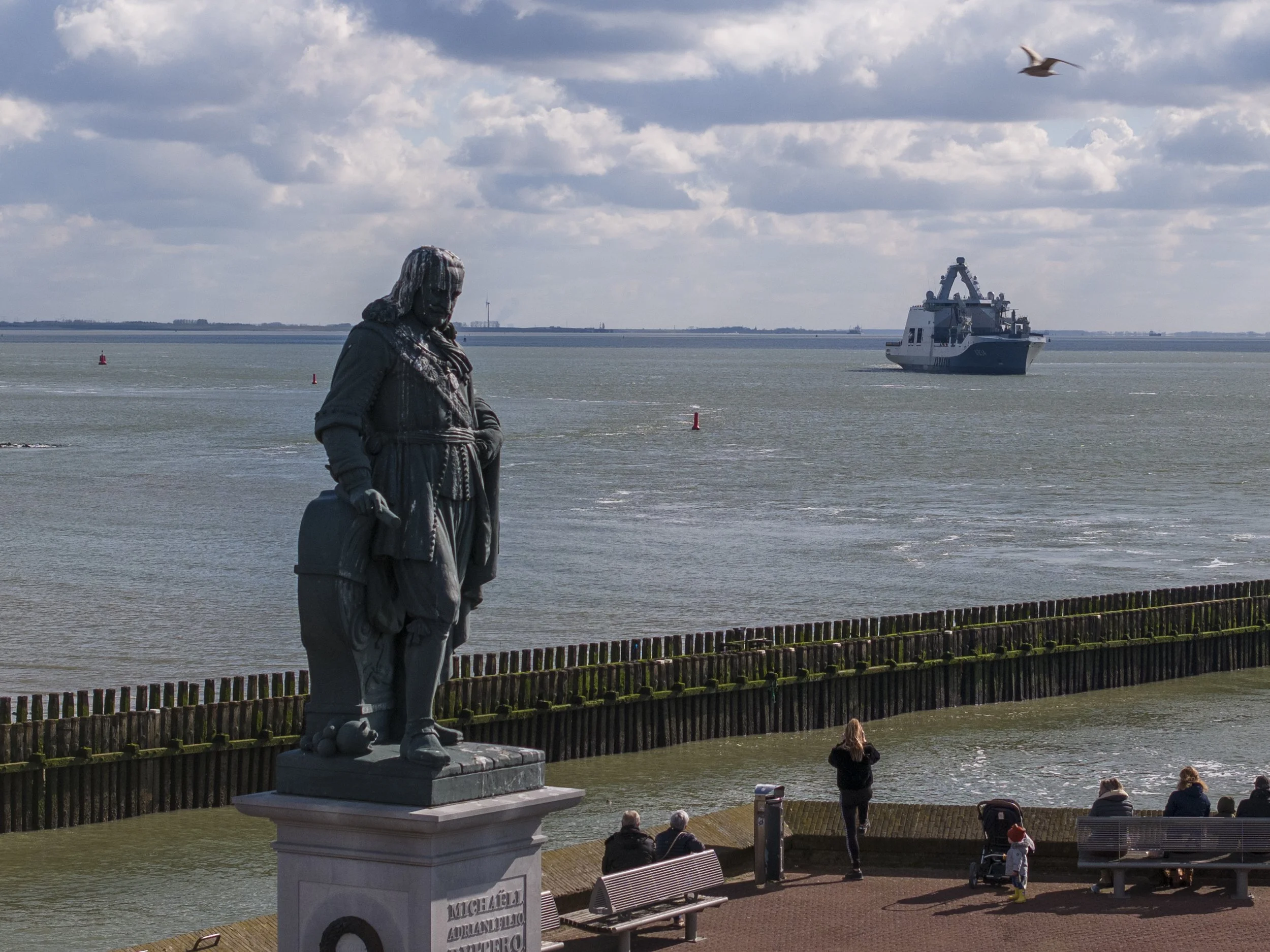 Statue of Michael Adriano with a suitcase, overlooking a harbor with a ship in the distance and people sitting on benches.
