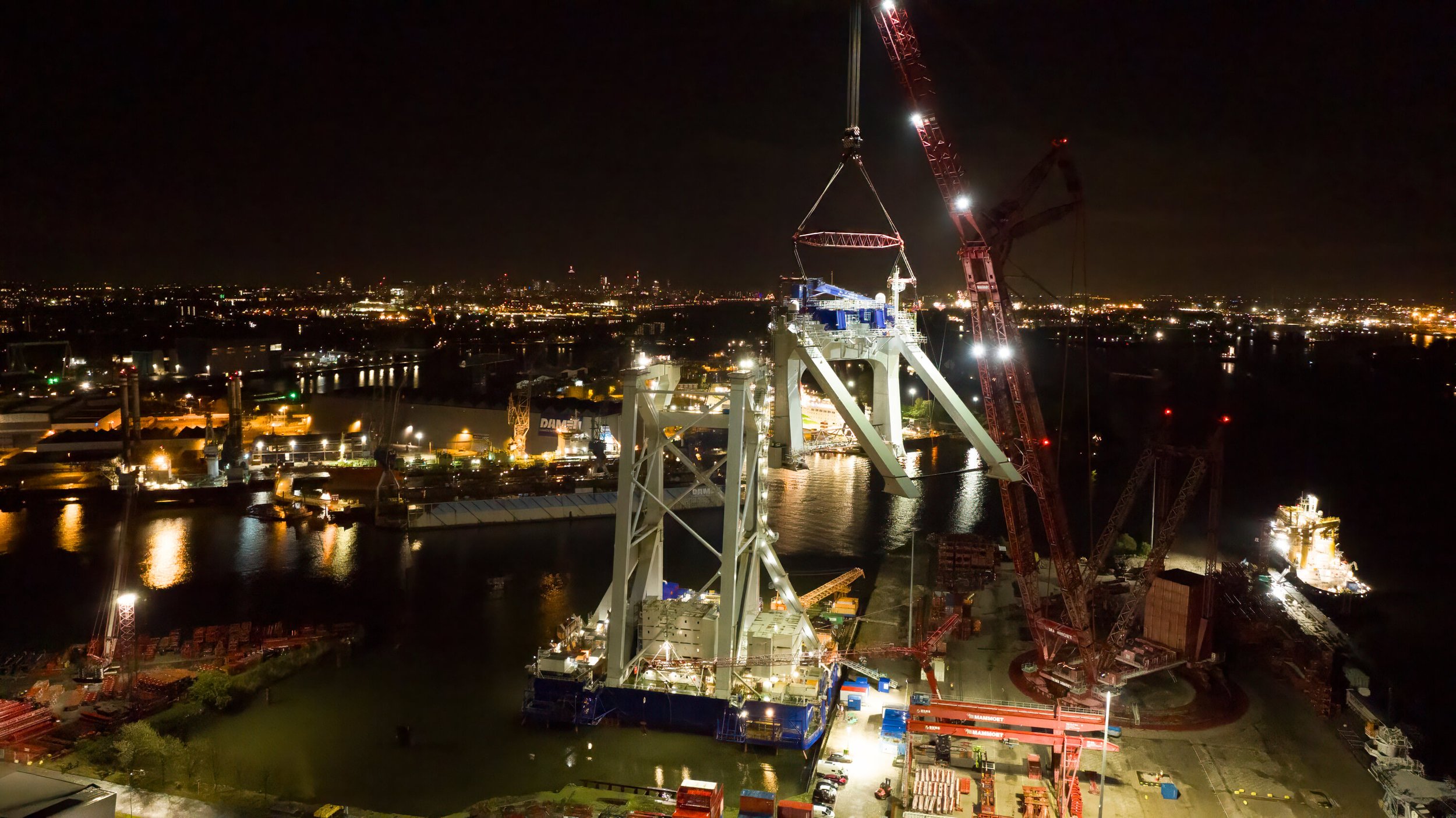 Nighttime construction site of a large bridge over water, with cranes and machinery illuminated, showcasing a partially built white bridge structure against a city skyline with lights in the distance.