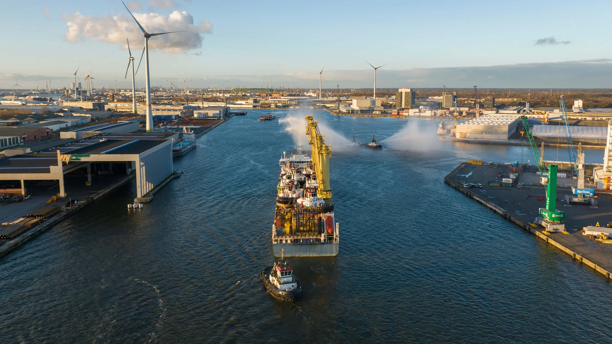 Aerial view of a busy port with a cargo ship being guided by tugboats, large cranes, warehouses, wind turbines, and industrial buildings, under a partly cloudy sky.
