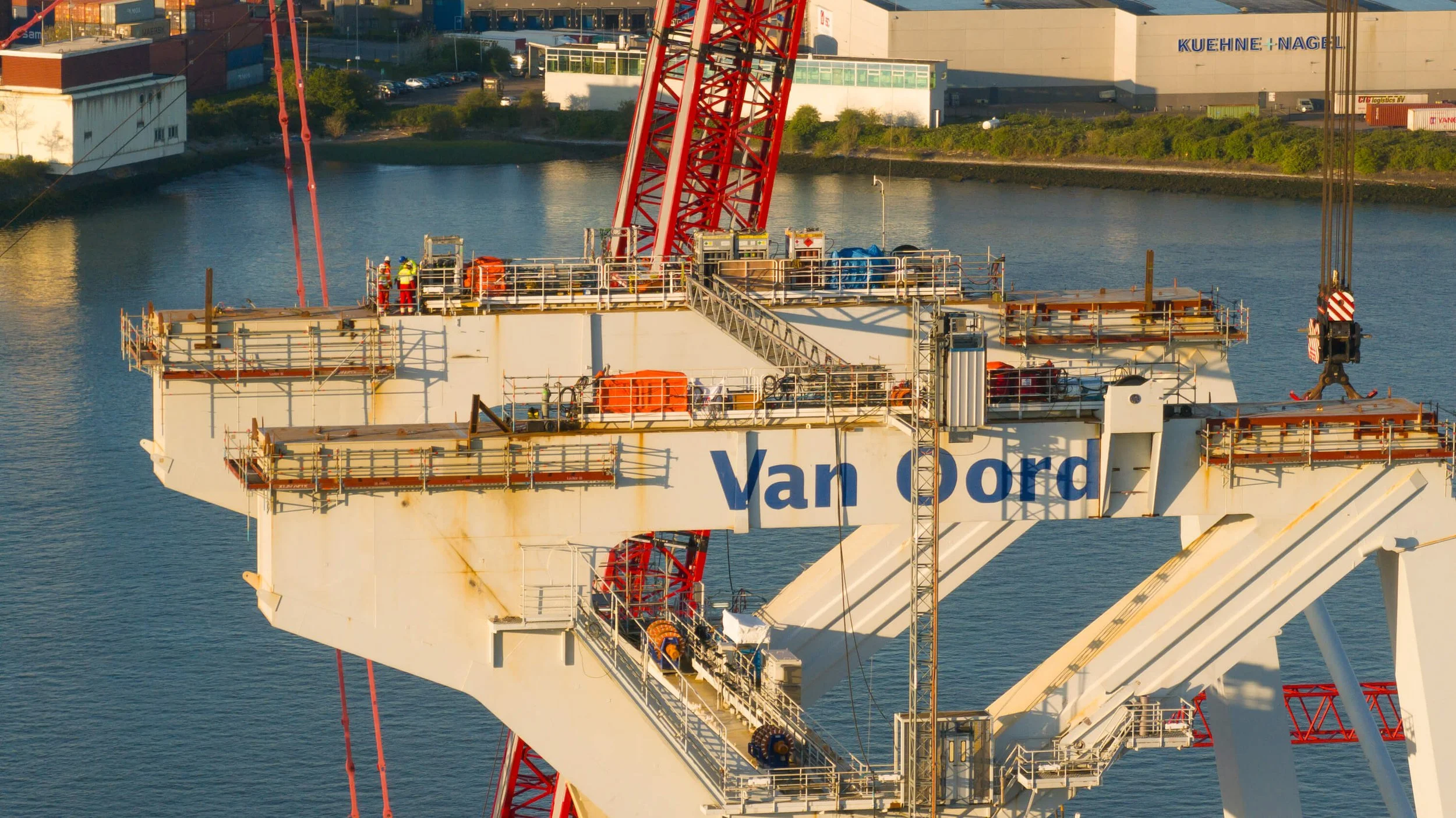 Close-up of the top section of a large offshore oil rig with workers, safety railings, and equipment, with water and industrial buildings in the background.