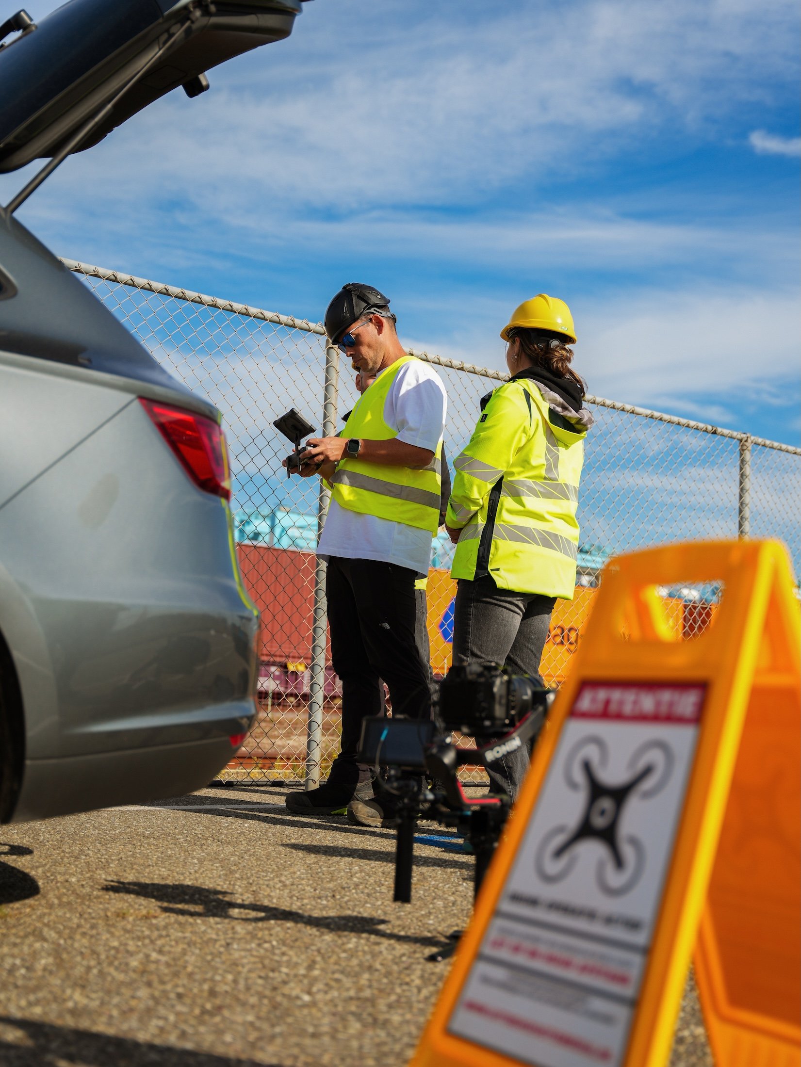 Two construction workers in high-visibility yellow jackets and helmets working near a gray car at a construction site, with warning signs and camera equipment on the ground.