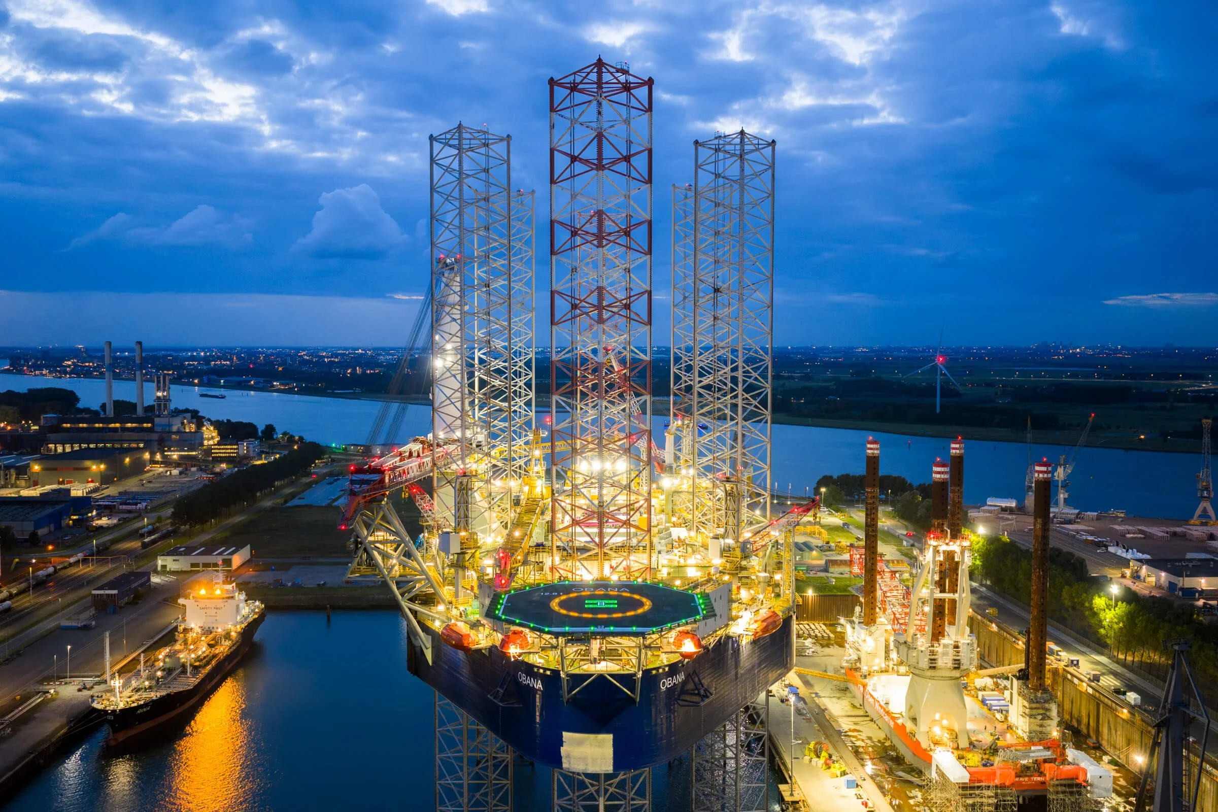 A large offshore oil rig illuminated with bright lights at dusk, with a helicopter landing pad on the platform and a river and cityscape in the background.