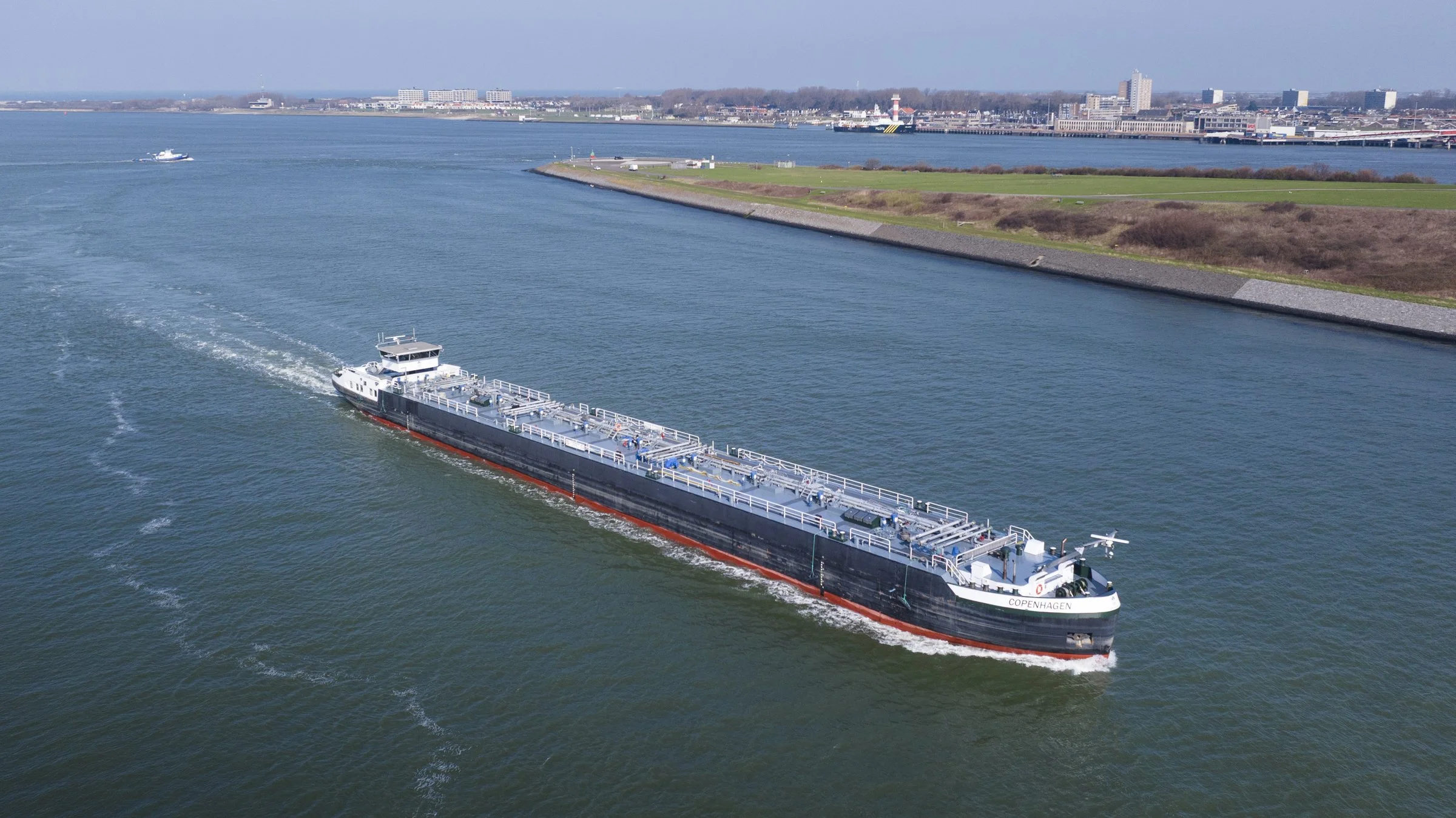 A large black and white cargo ship named Copenhagen is sailing through a harbor, with a city skyline and shoreline visible in the background.