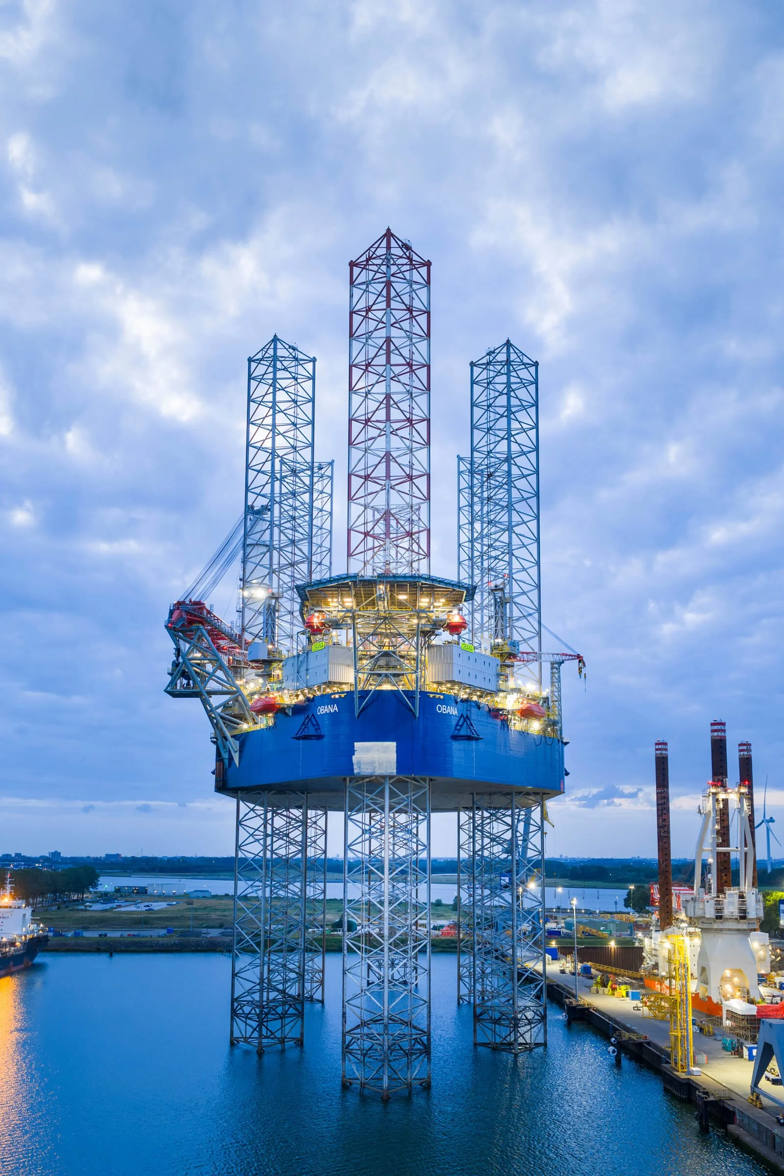 Offshore oil drilling platform with tall structure, situated in water, under a cloudy sky at dusk.
