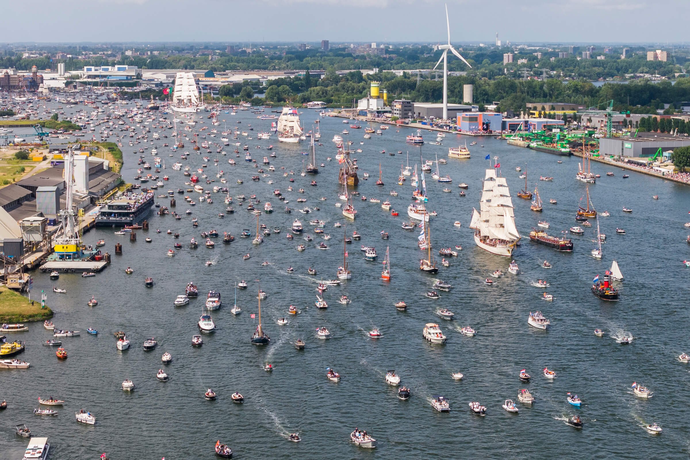 Aerial view of a harbor filled with numerous boats and sailing ships, with a city skyline and green trees in the background.