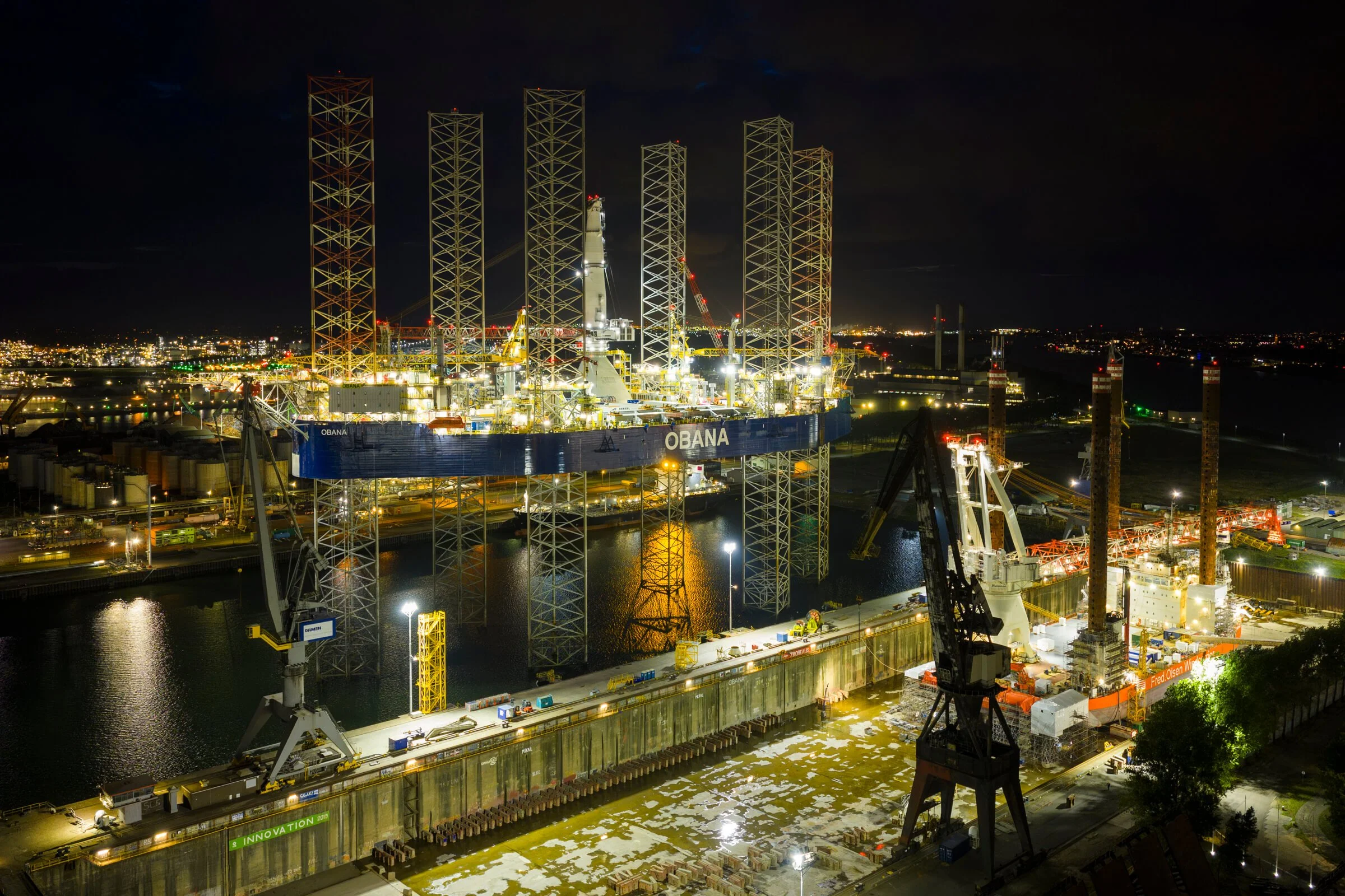 Nighttime view of an offshore oil rig with illuminated structures and cranes, surrounded by water and city lights in the background.