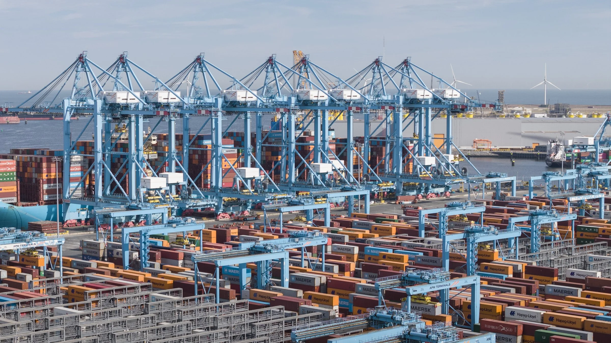 Cargo port with numerous shipping containers and large blue cranes, with wind turbines in the background.