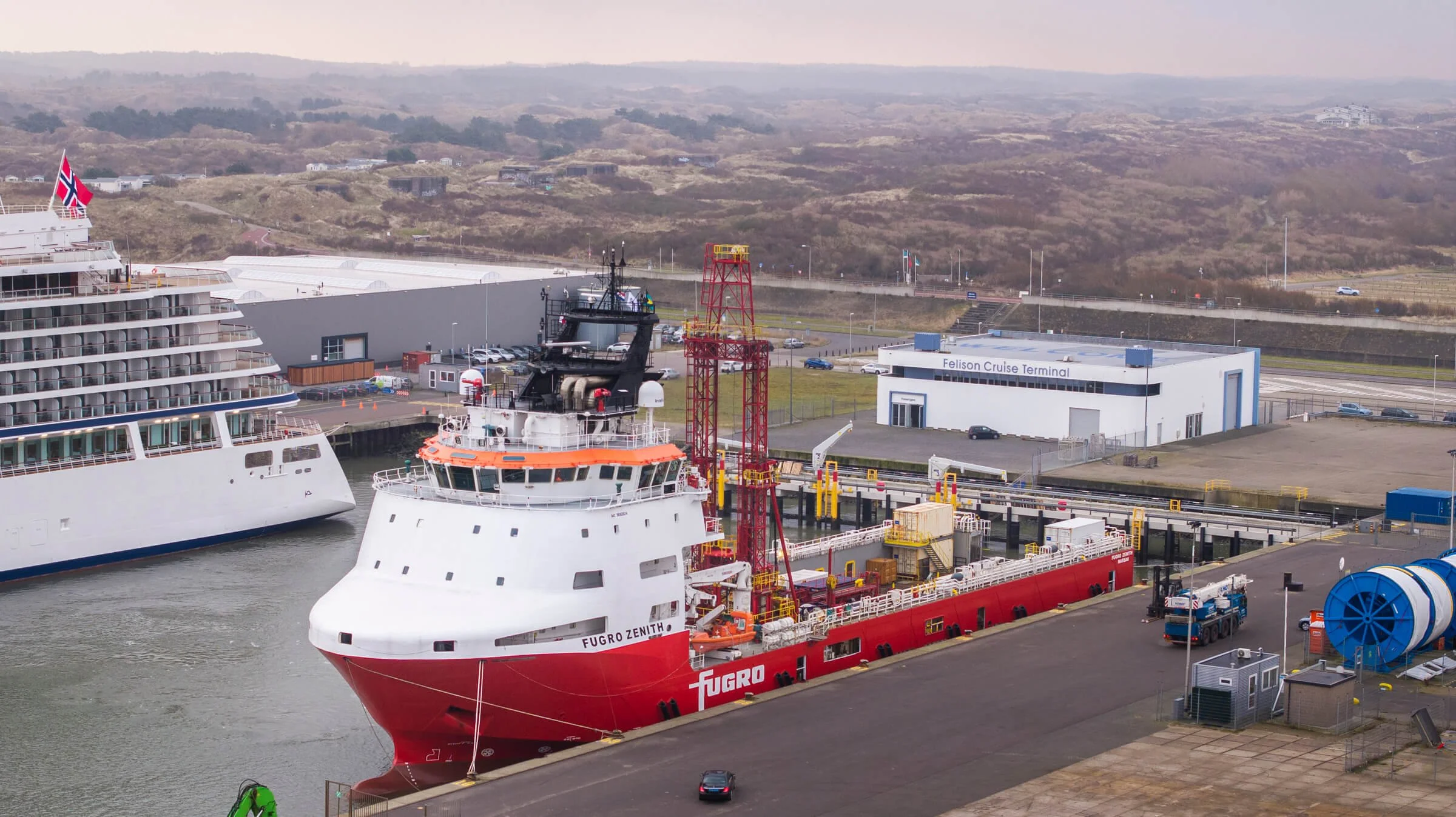 A large harbor scene featuring a red and white Fugro survey ship docked at a port, with other ships visible nearby. The harbor has industrial equipment, a building labeled Felison Cruise Terminal, and hilly terrain in the background.