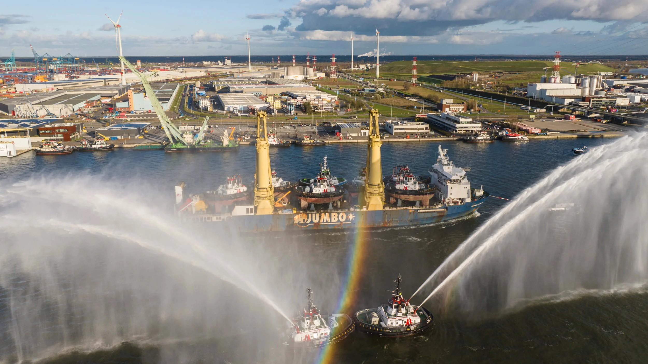 A cargo ship named Jumbo is being washed by two fireboats with water hoses, creating a rainbow in the spray, with an industrial port and wind turbines in the background.