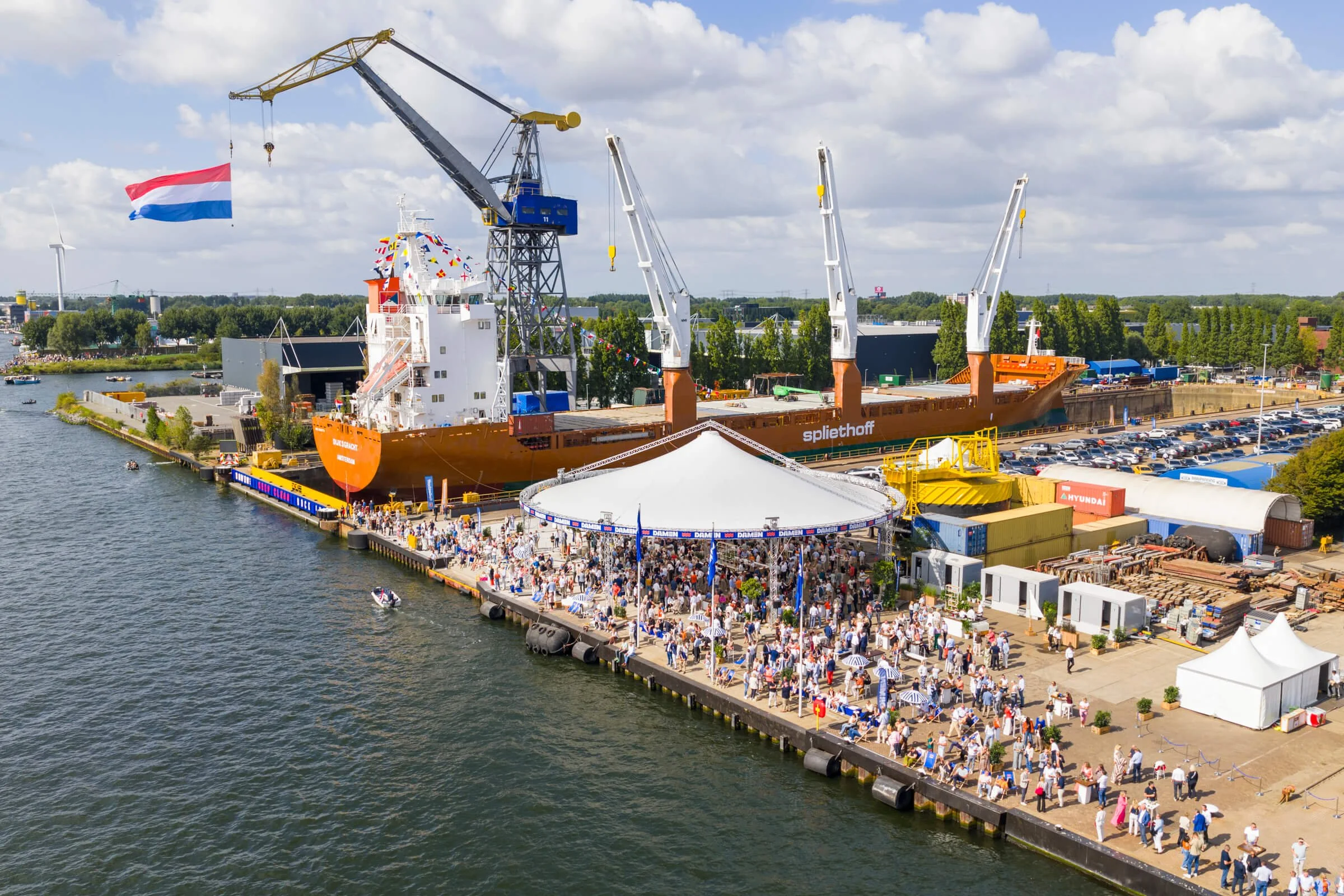 A large ship docked at a port with a crowd gathered on the pier and a white tent. The ship is decorated with colorful banners and has the name "Bunschoten" on the bow. A Dutch flag waves from the ship. Several cranes are adjacent to the ship, and the