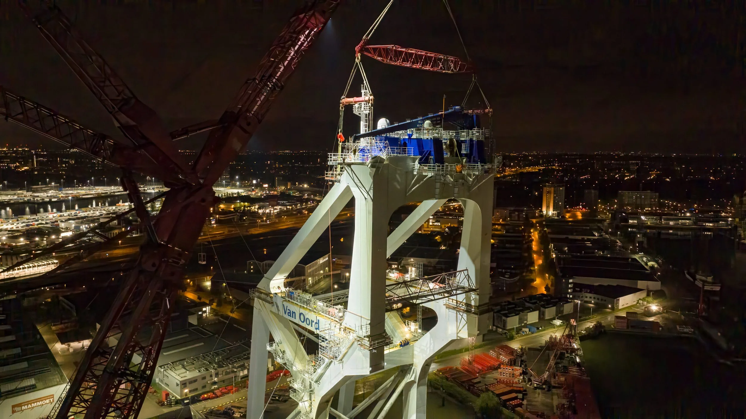Nighttime view of a large construction crane and a tall bridge structure with a cityscape in the background, illuminated by street and building lights.