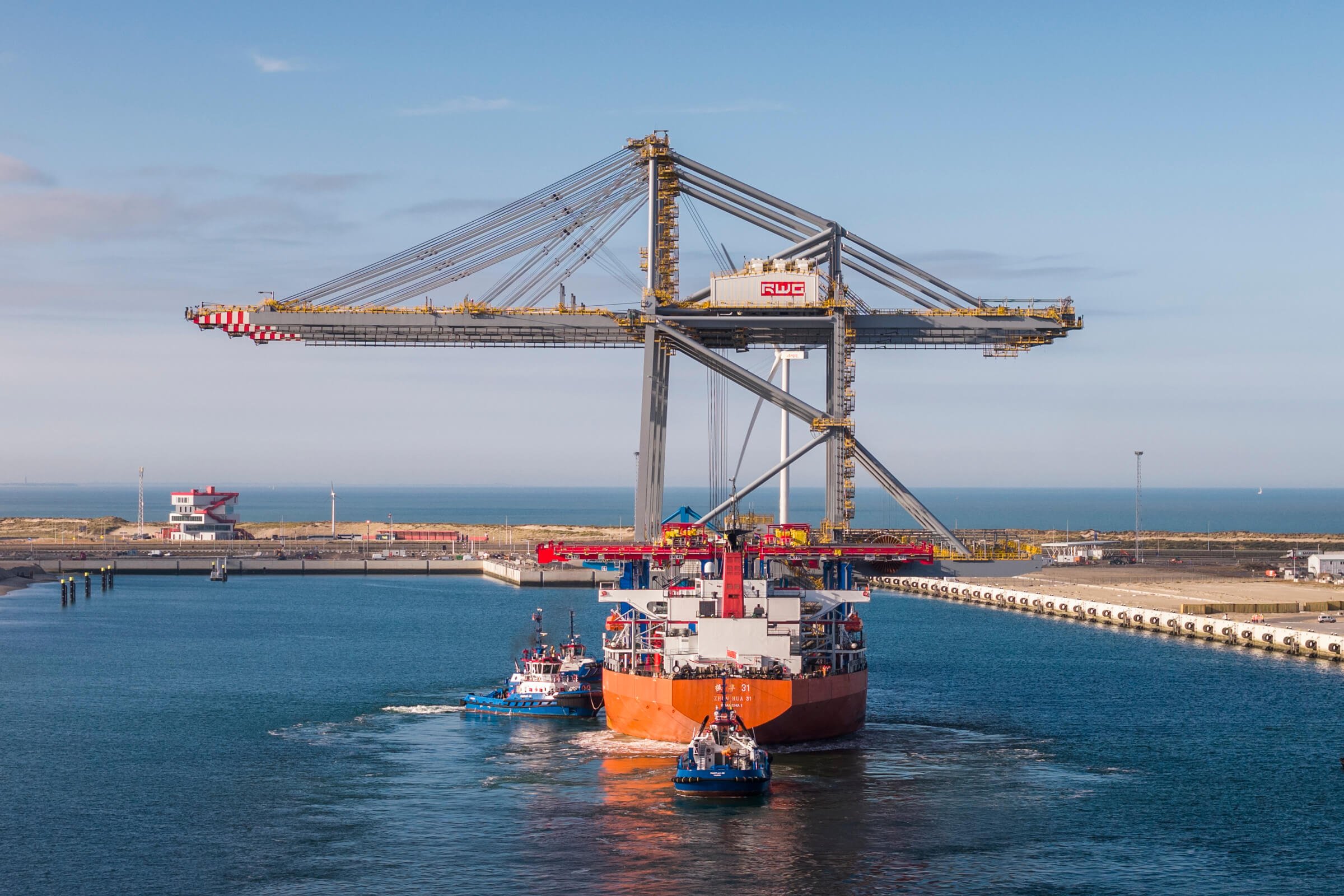Large cargo ship being guided through a harbor by two tugboats, with a massive port crane in the background and water on either side.