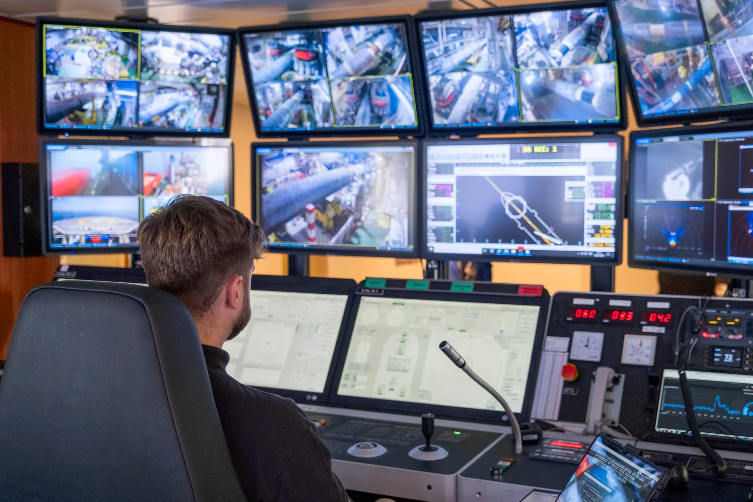 A man sitting in a control room with multiple computer monitors displaying surveillance footage and technical data, possibly for security or industrial monitoring.