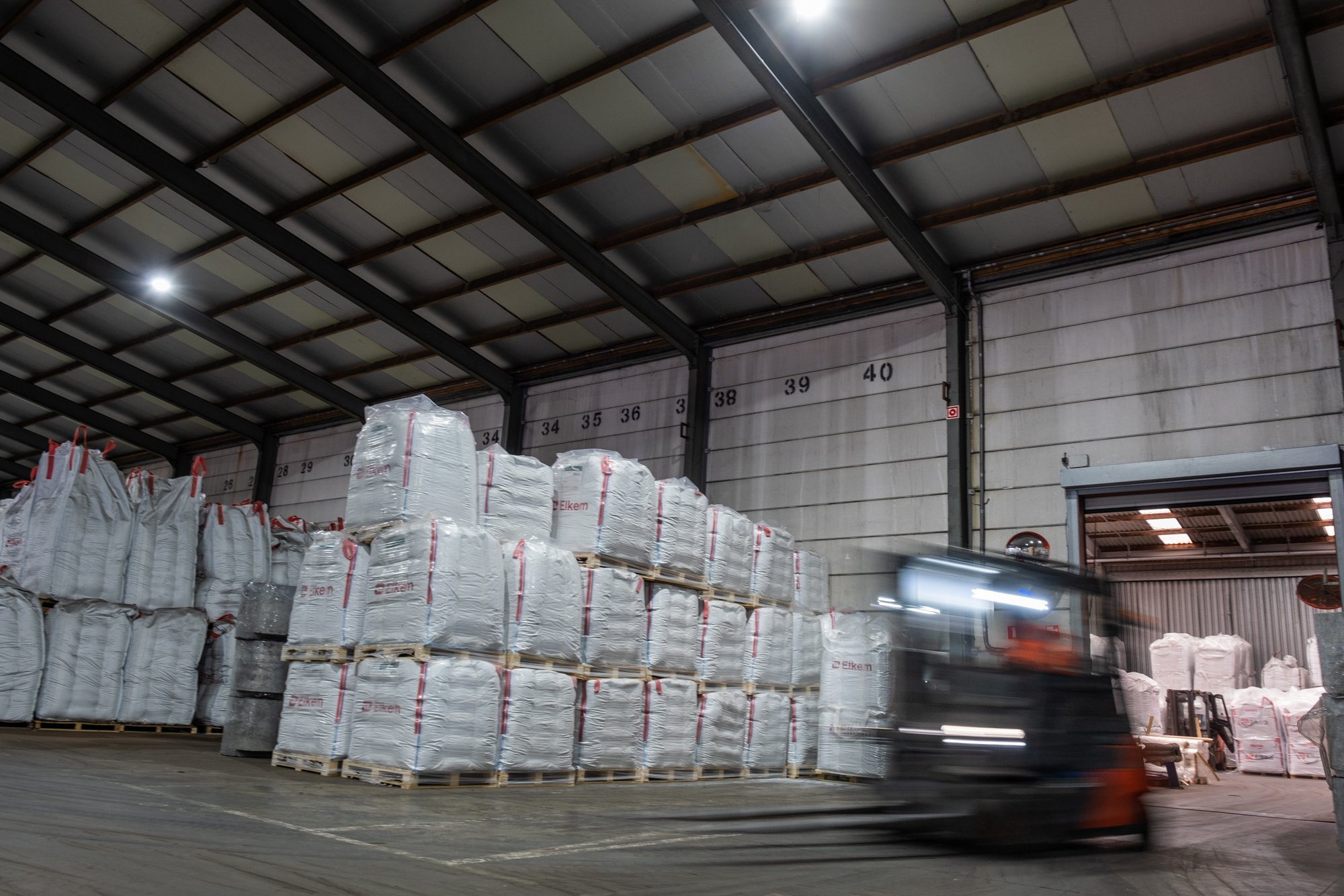 A warehouse with stacks of wrapped pallets and bags, a forklift moving quickly, high ceiling, and numbered storage bays