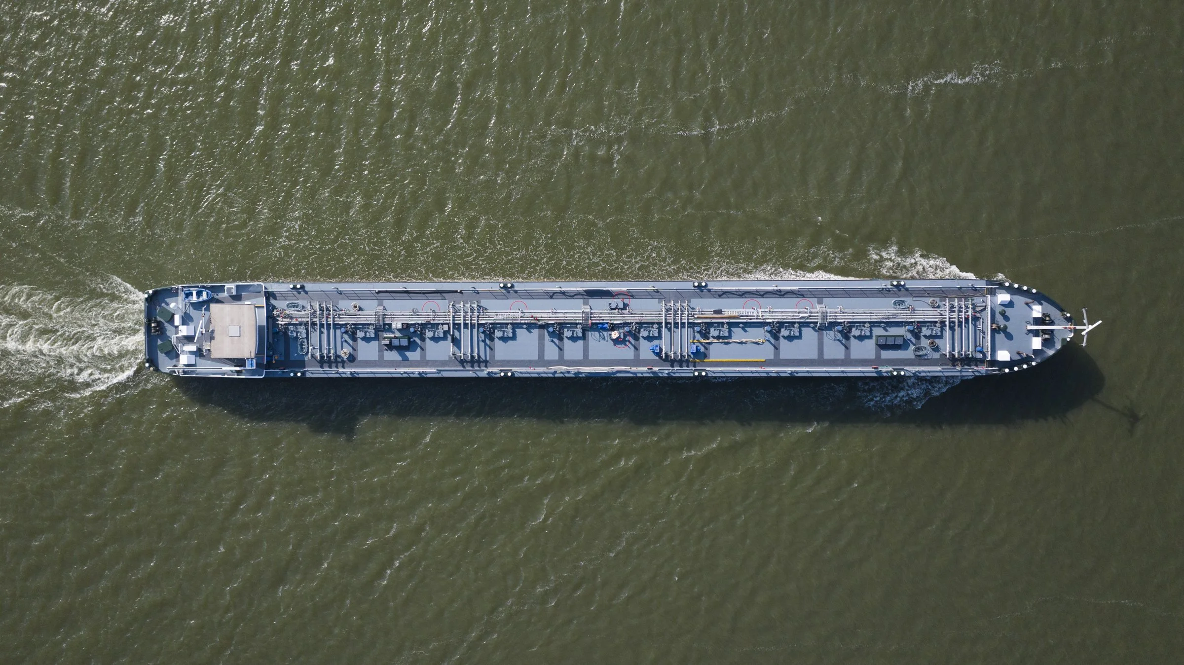 An aerial view of a large gray cargo ship sailing through greenish water, creating waves behind it.
