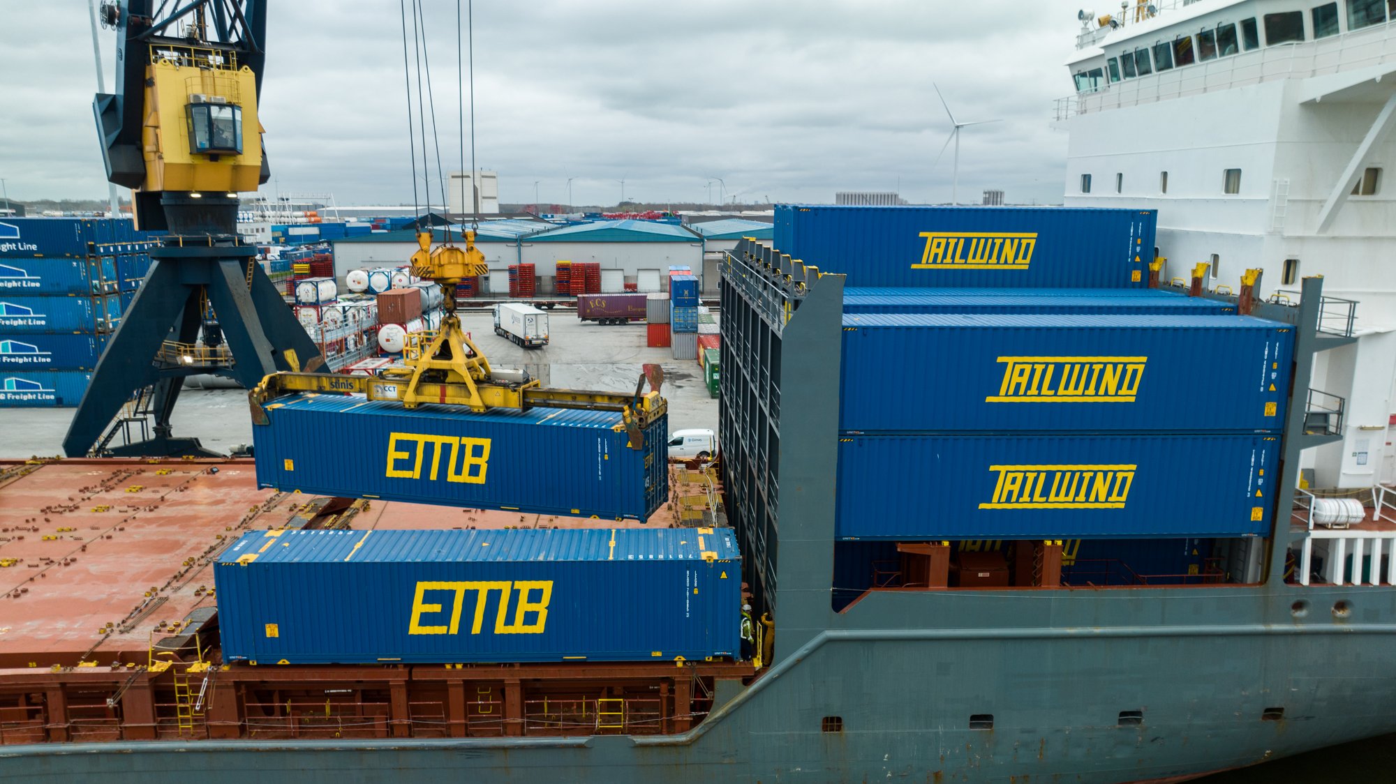 A cargo ship at port loading large blue shipping containers with yellow lettering, using a crane for lifting containers onto the ship.
