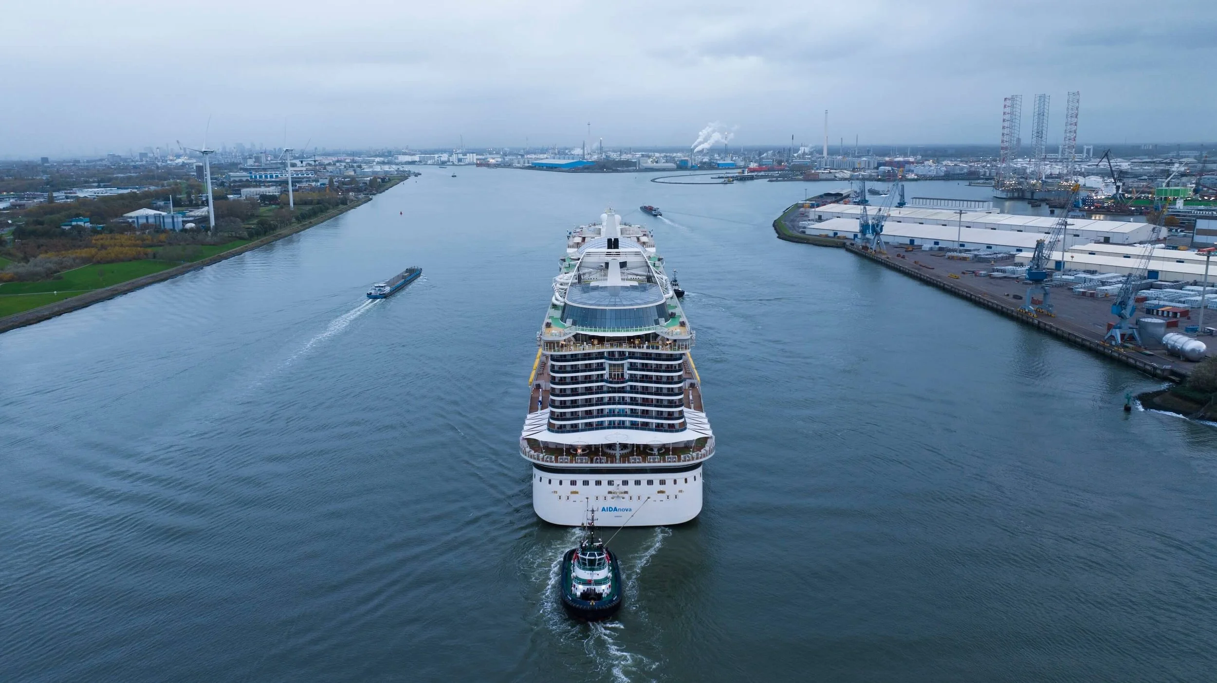A large cruise ship being guided through a canal by a small tugboat, with industrial port facilities on the right and a city skyline with wind turbines in the background.