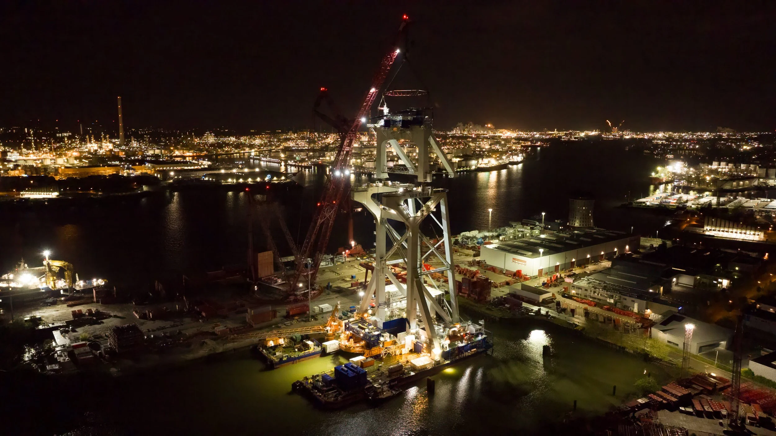 Nighttime construction site by the water with large cranes, industrial buildings, and city lights in the background.