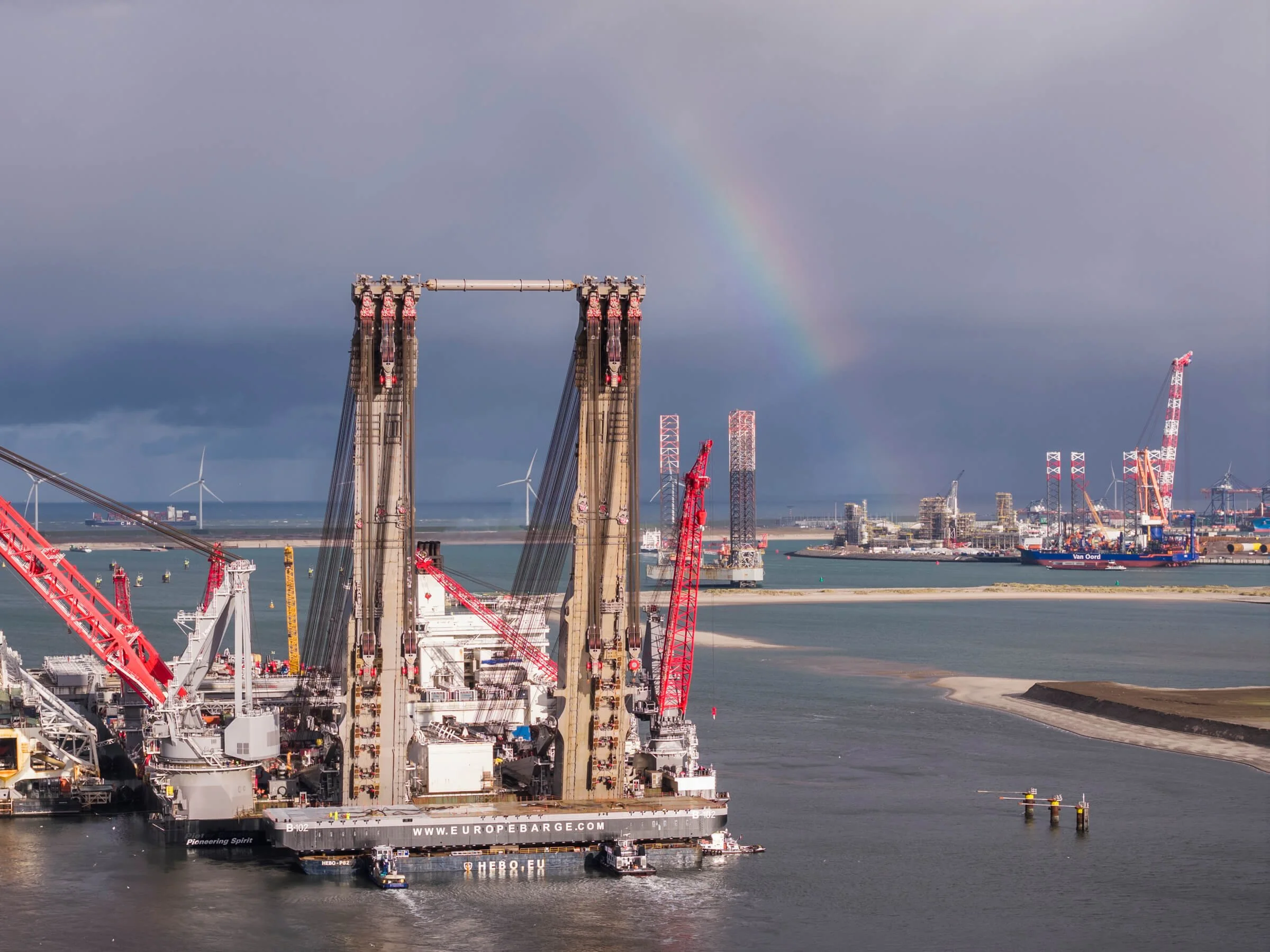Large construction barges with cranes on a body of water, with wind turbines and harbor in the background, a rainbow in the cloudy sky.