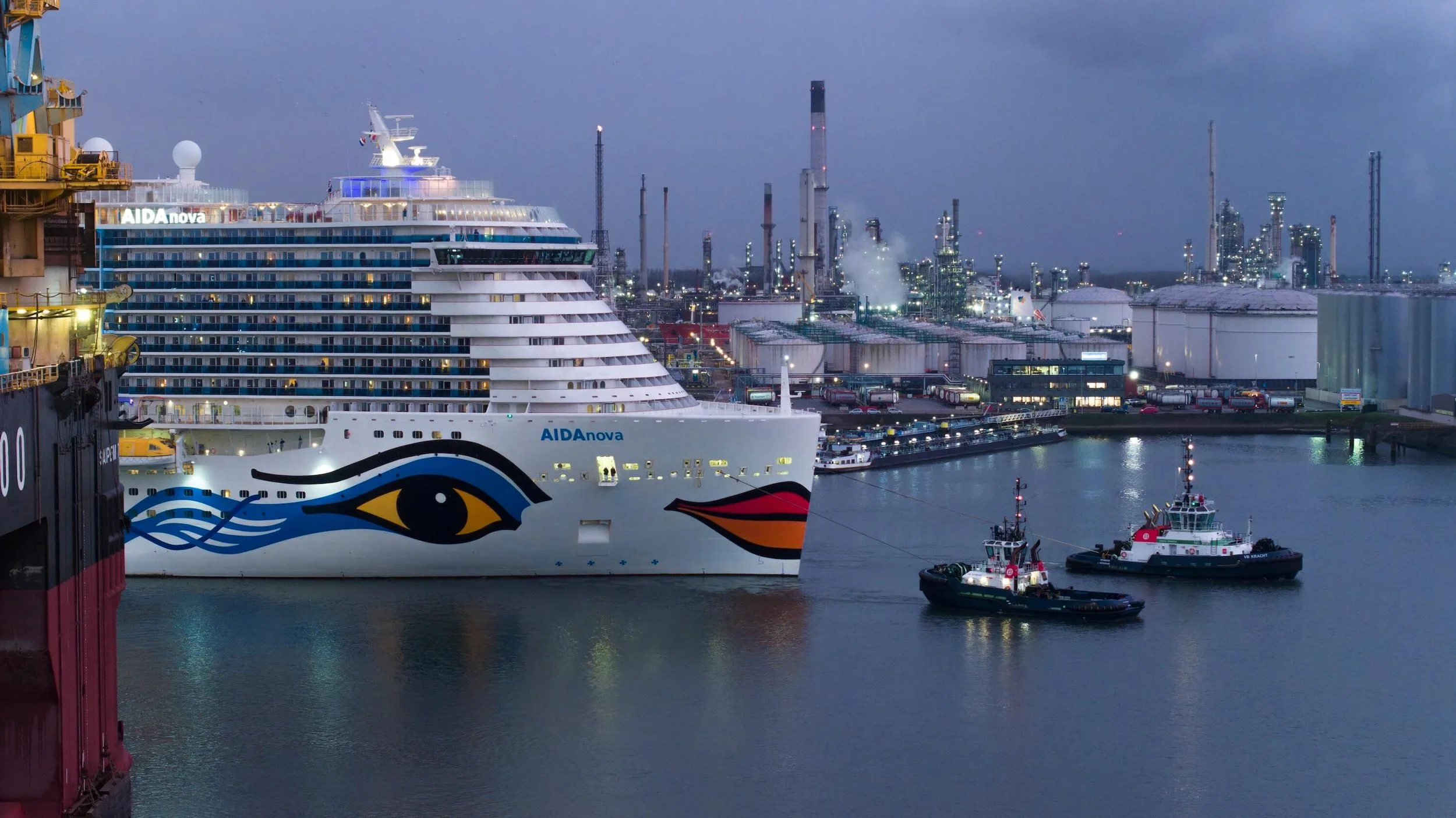 A large cruise ship named AIDAnova docked at a port with two tugboats near it, industrial buildings and factories in the background, and a city skyline visible in the distance during dusk.