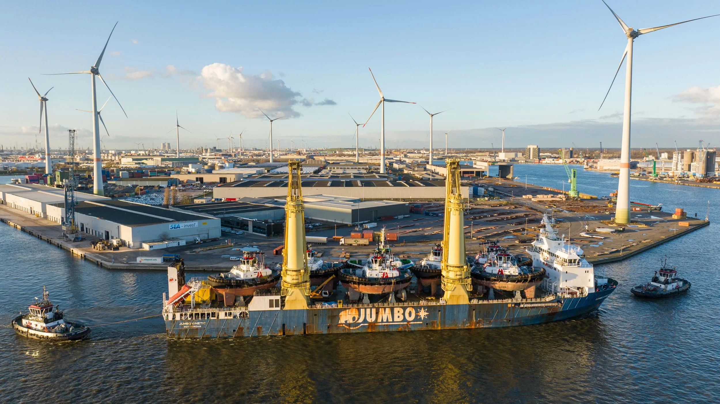 Cargo ship named Jumbo with multiple boats on top, docked at a port with wind turbines and industrial buildings, under a blue sky with some clouds.