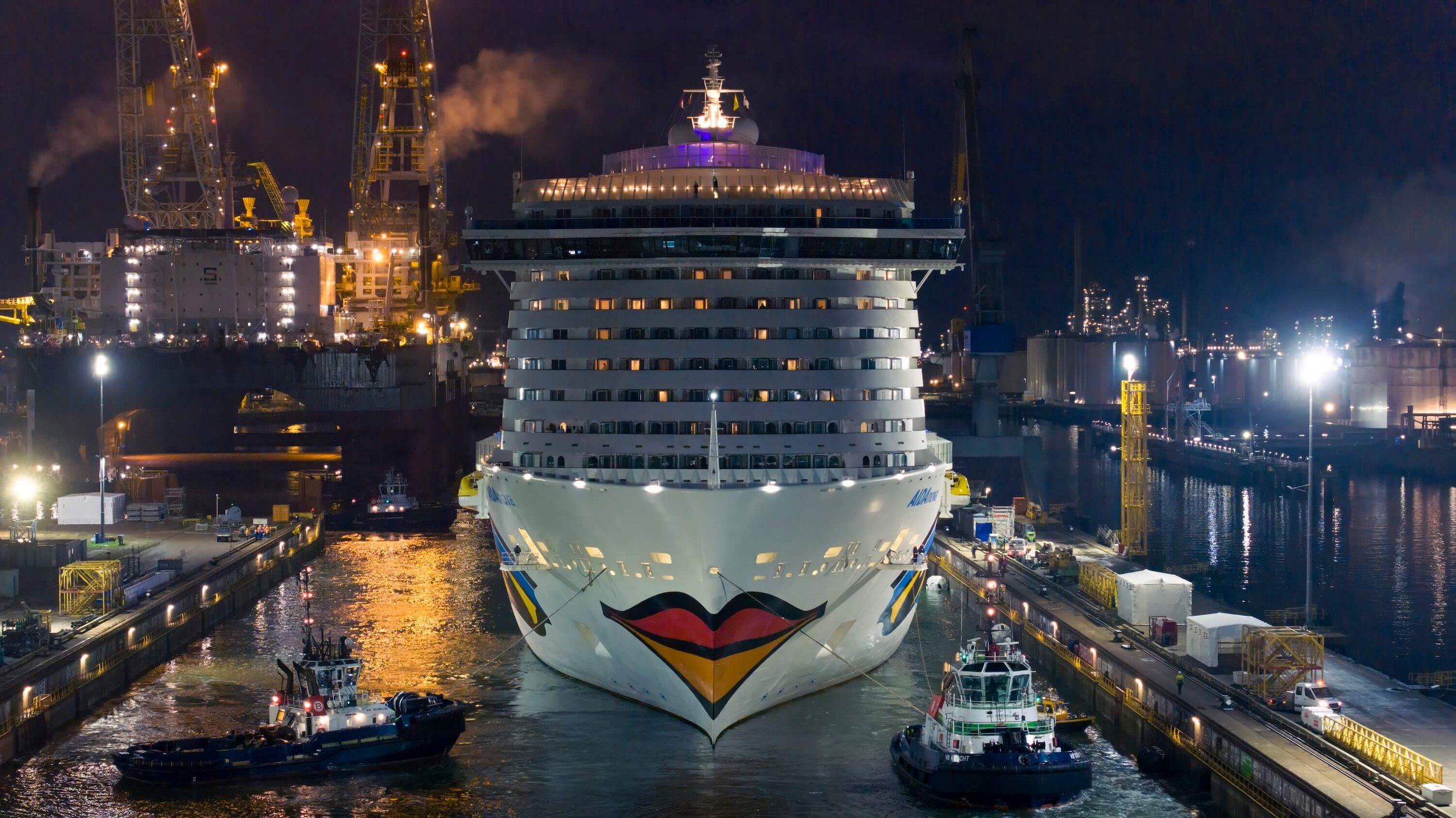 A large cruise ship docked at a port during nighttime, with bright lights illuminating the ship and the surrounding dock area, and industrial infrastructure in the background.