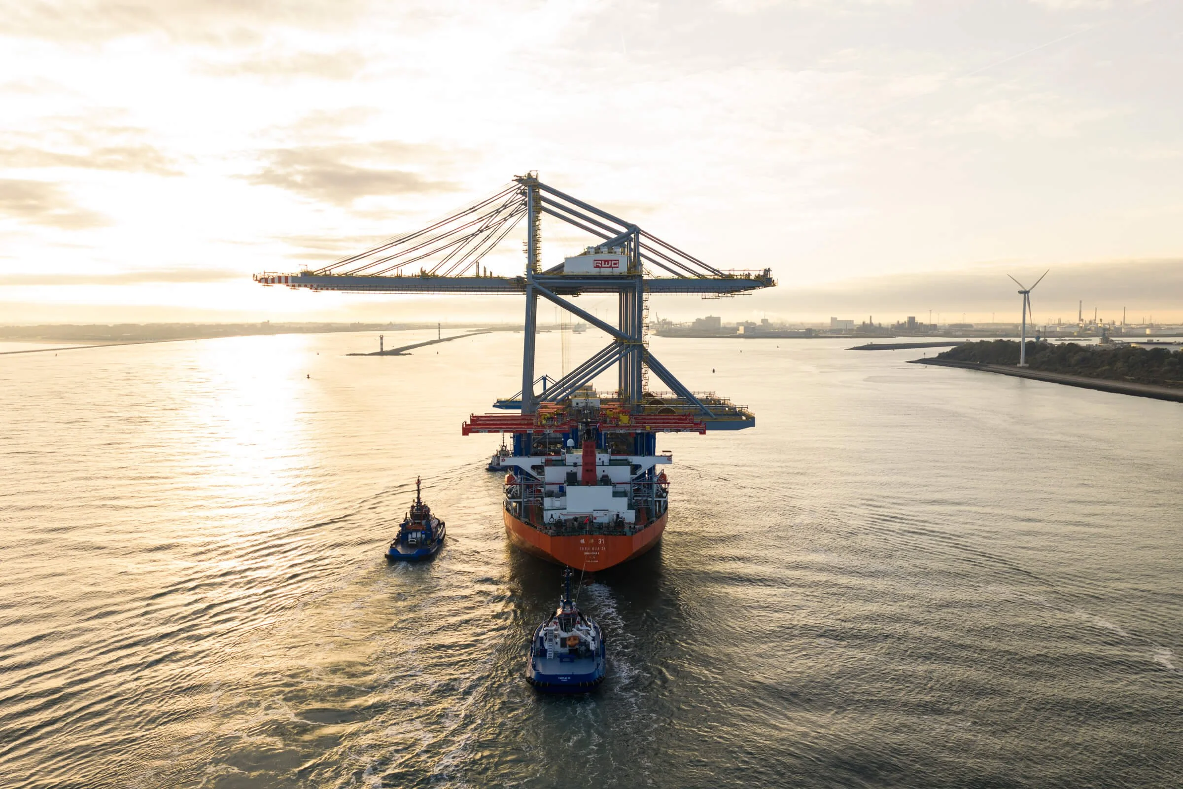 A large cargo ship being maneuvered through a harbor by three tugboats with a sunset sky and wind turbines visible in the distance.