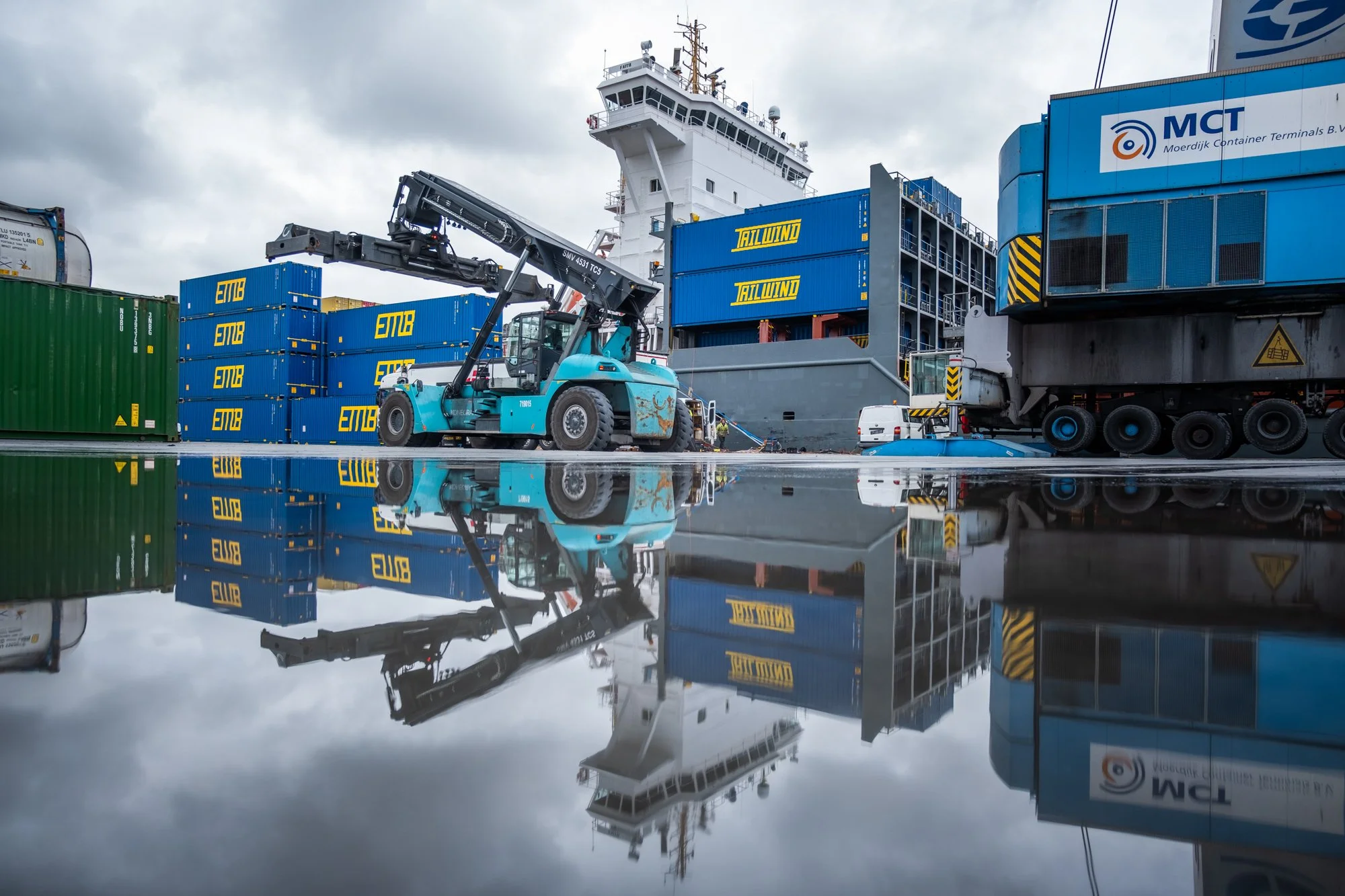 View of a container terminal with stacked shipping containers, a forklift, and a cargo ship, reflected in a puddle of water on the ground.