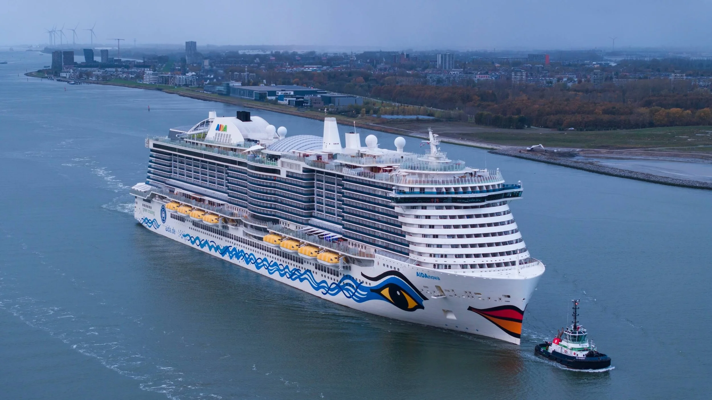A large cruise ship named AIDAnova with a colorful eye and lips design on its hull, leaving a port and being guided by a smaller tugboat in the water with an urban cityscape and wind turbines in the background.
