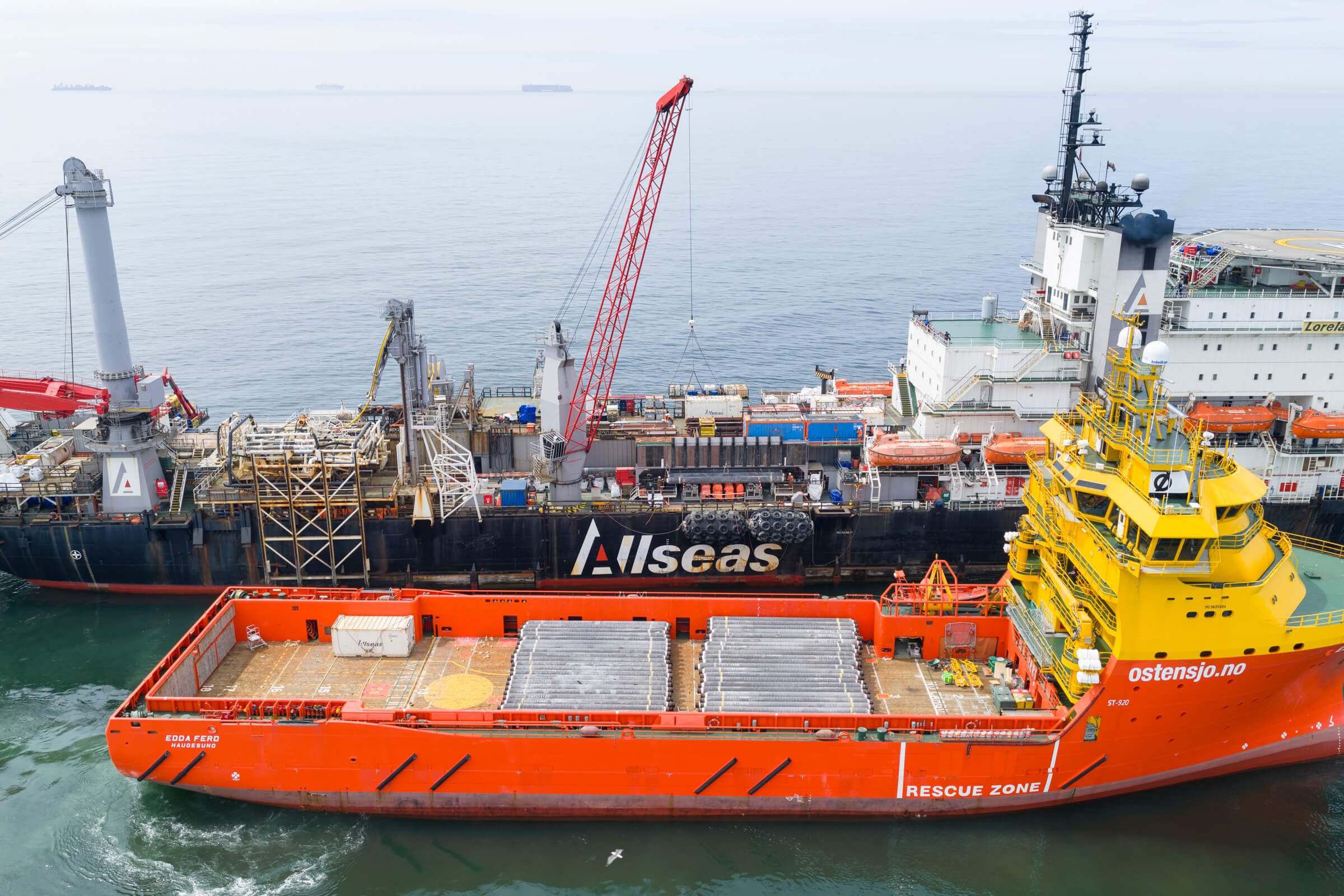 A large orange rescue ship with the words 'Rescue Zone' on its side, docked beside a black oil supply vessel with 'Alsas' branding, in a harbor with calm water and several ships in the background.