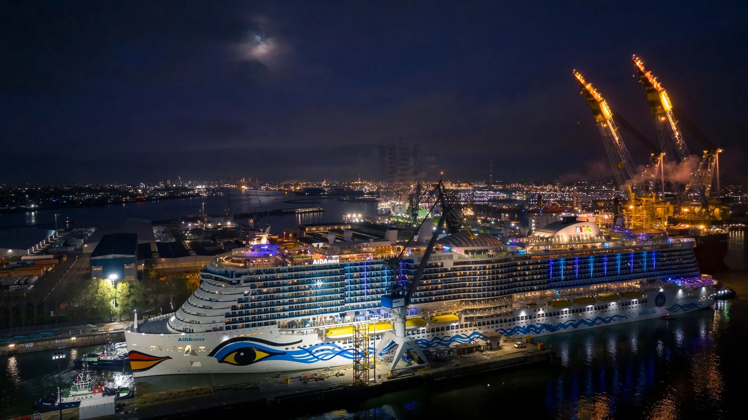 A large illuminated cruise ship named AIDAnova docked at a port at night, with cranes and city lights in the background, and a partly visible moon in the sky.