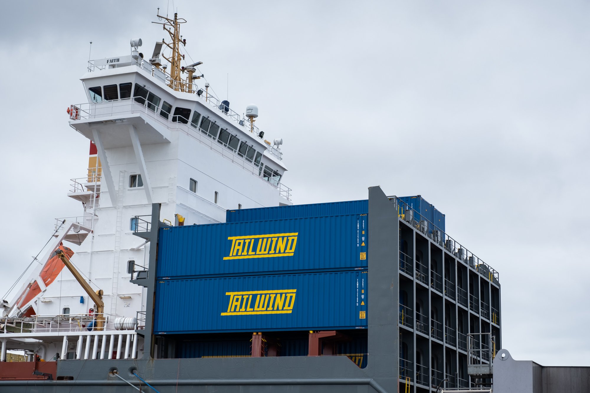Close-up of a cargo ship's bridge and stacked shipping containers with the logo 'TALWIND' in yellow, under a cloudy sky.
