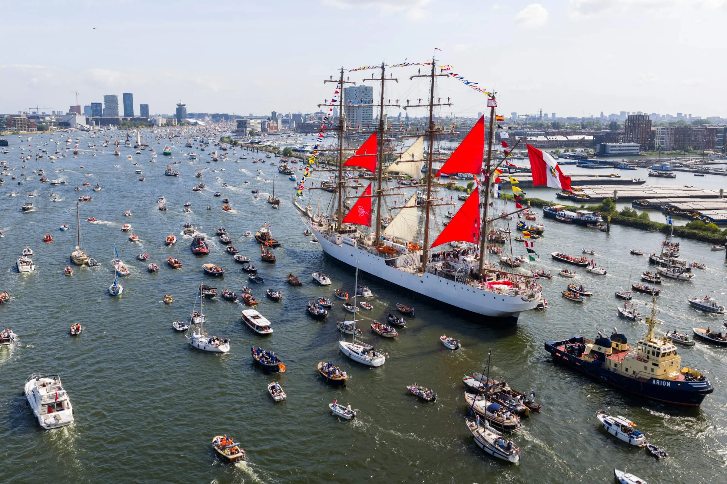 A large sailing ship with red sails and pennants, surrounded by numerous smaller boats on a river in an urban area, with city buildings in the background.