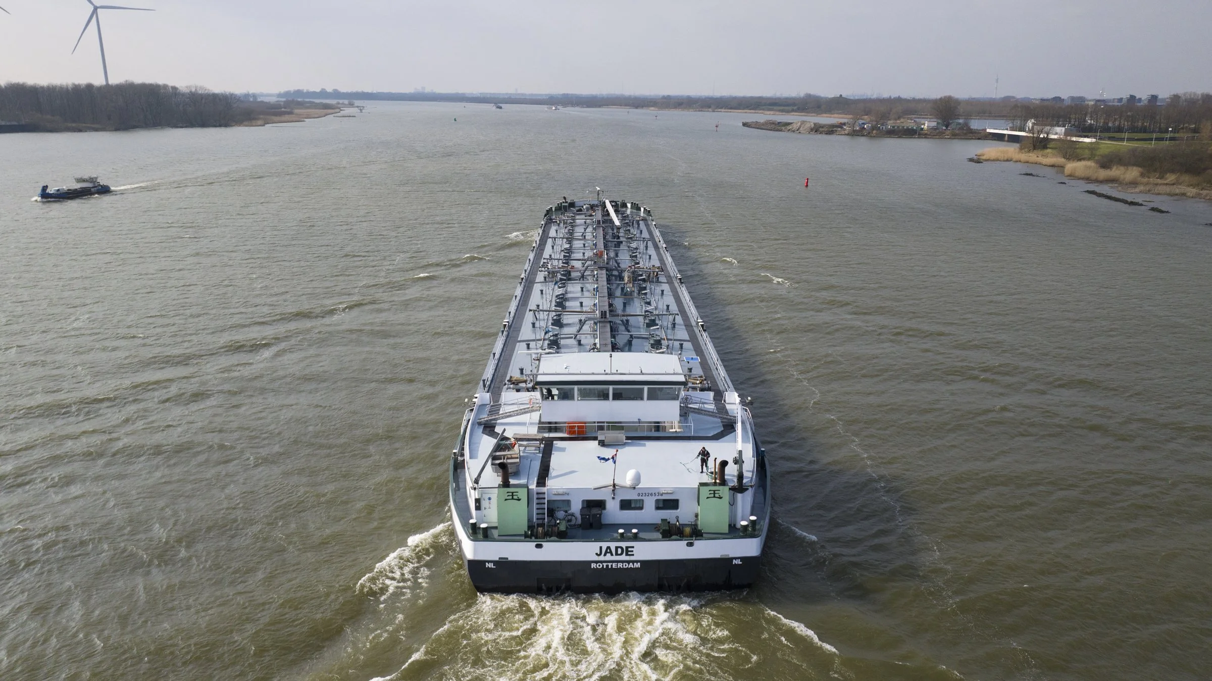 A large cargo ship named Jade traveling on a wide river with a smaller boat in the background. The ship is heading away from the camera, creating waves at the stern.