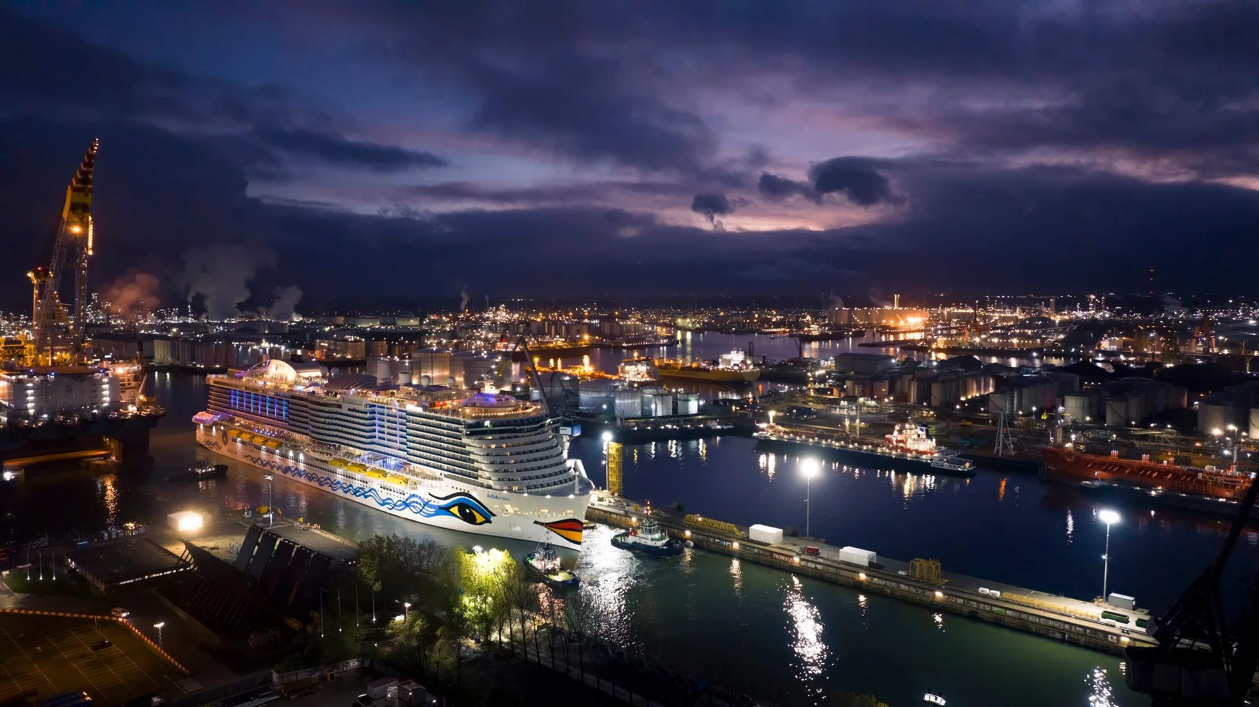 Night view of a busy harbor with a large cruise ship decorated with an eye and wave patterns, illuminated against a dark sky, surrounded by industrial buildings, cranes, and city lights reflecting on the water.