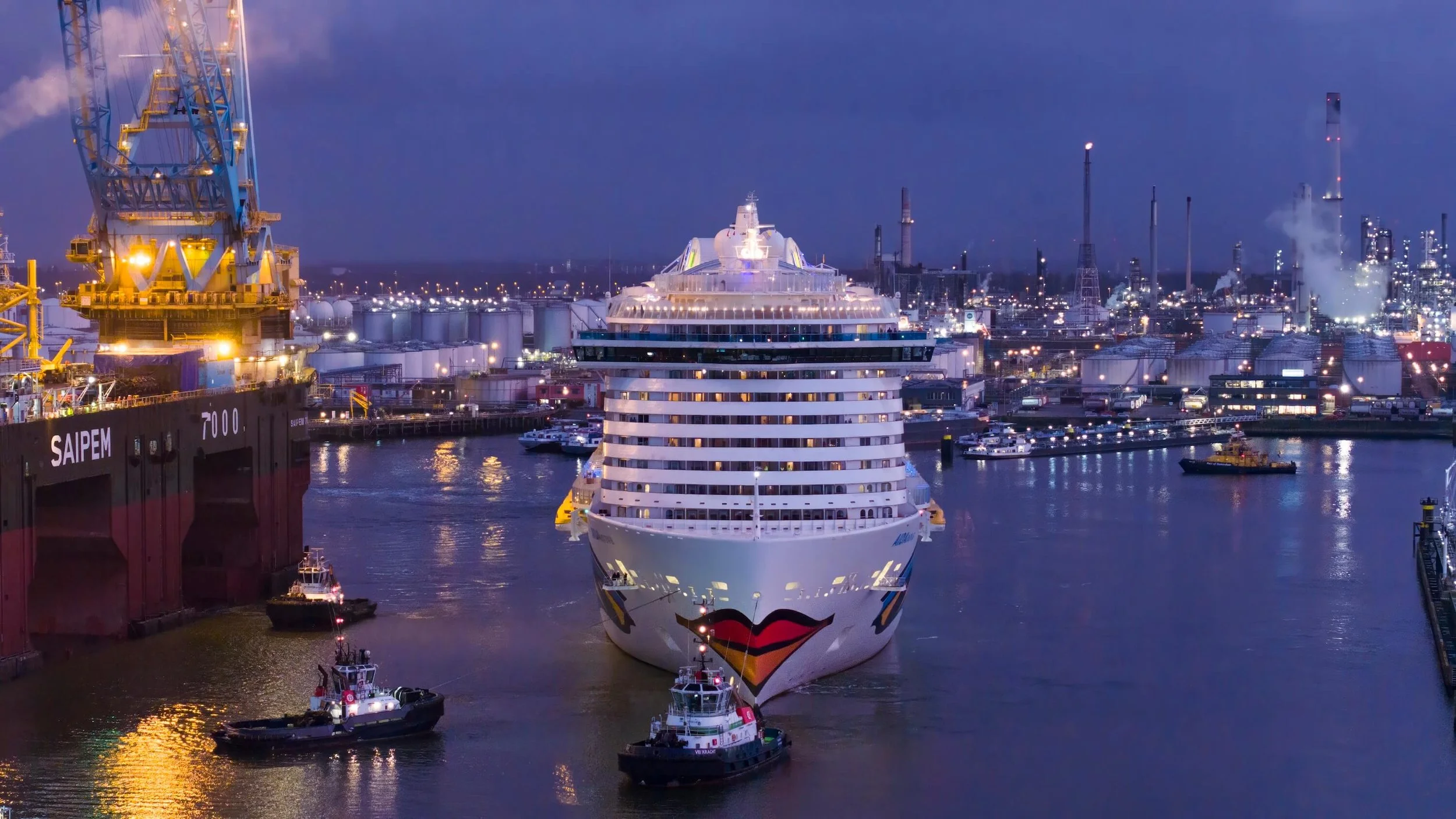 Cruise ship with colorful lip design on the front, arriving at a port with tugboats during evening on a cloudy night, surrounded by industrial buildings and other boats.